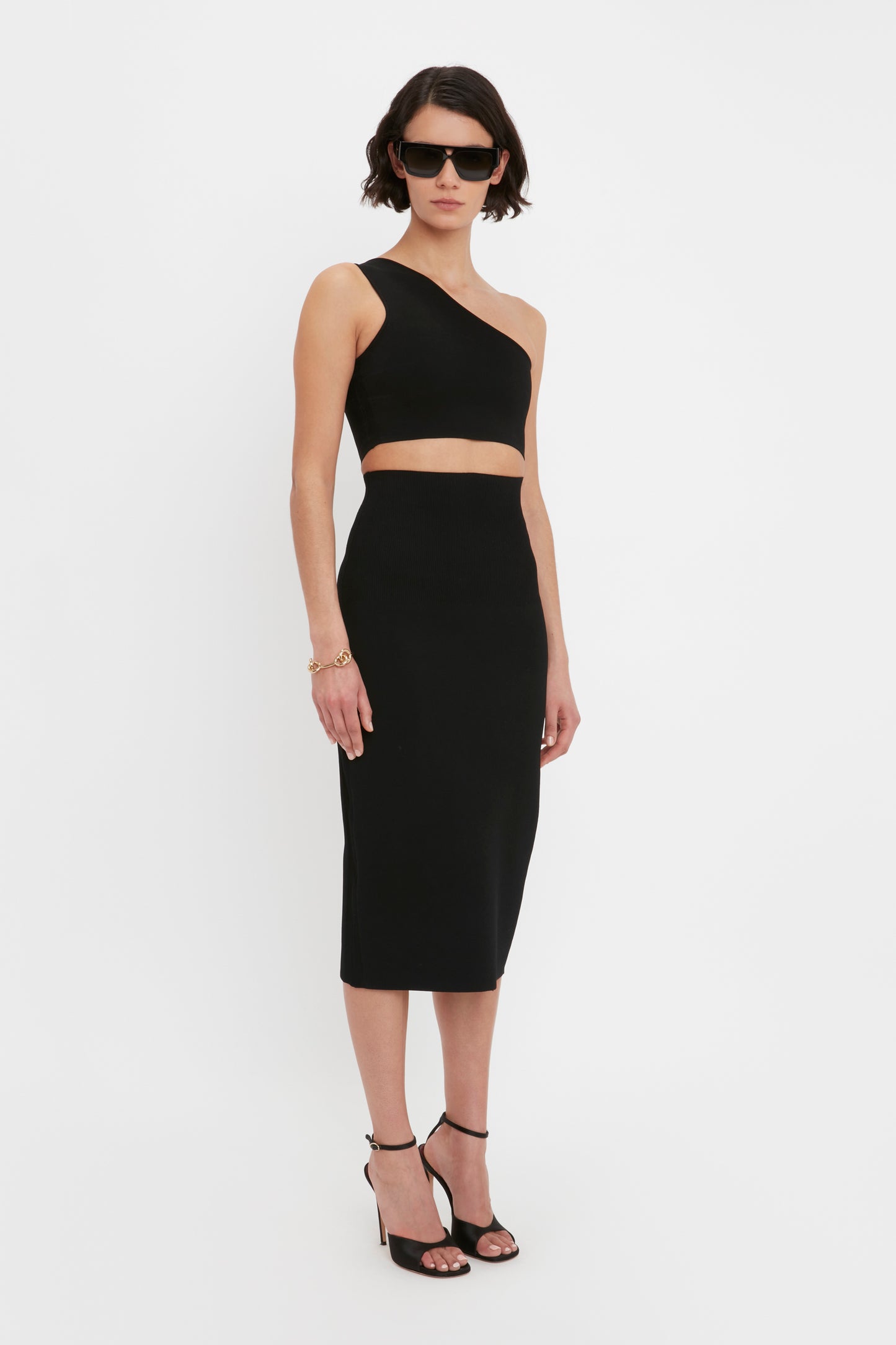 A person with short dark hair wearing sunglasses, the VB Body One Shoulder Crop Top by Victoria Beckham in black, and a matching black midi skirt with high heels, standing against a plain white background.