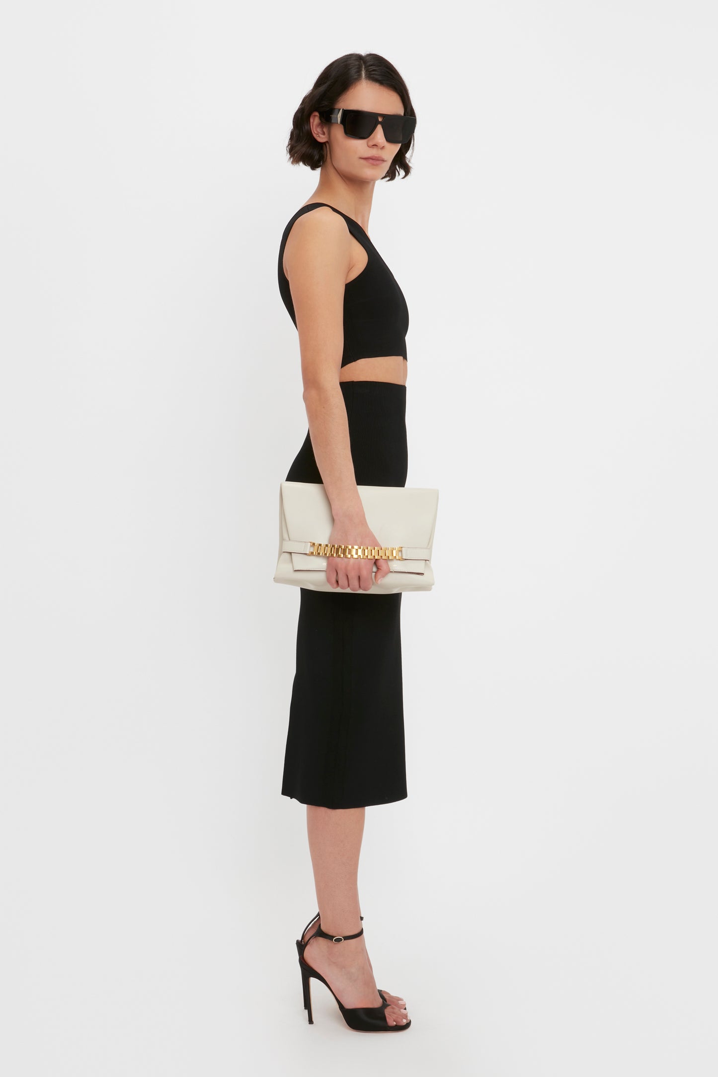 Person wearing a black sleeveless top, a VB Body Fitted Midi Skirt in Black by Victoria Beckham, high heels, and sunglasses, holding a beige clutch with a gold chain accent.