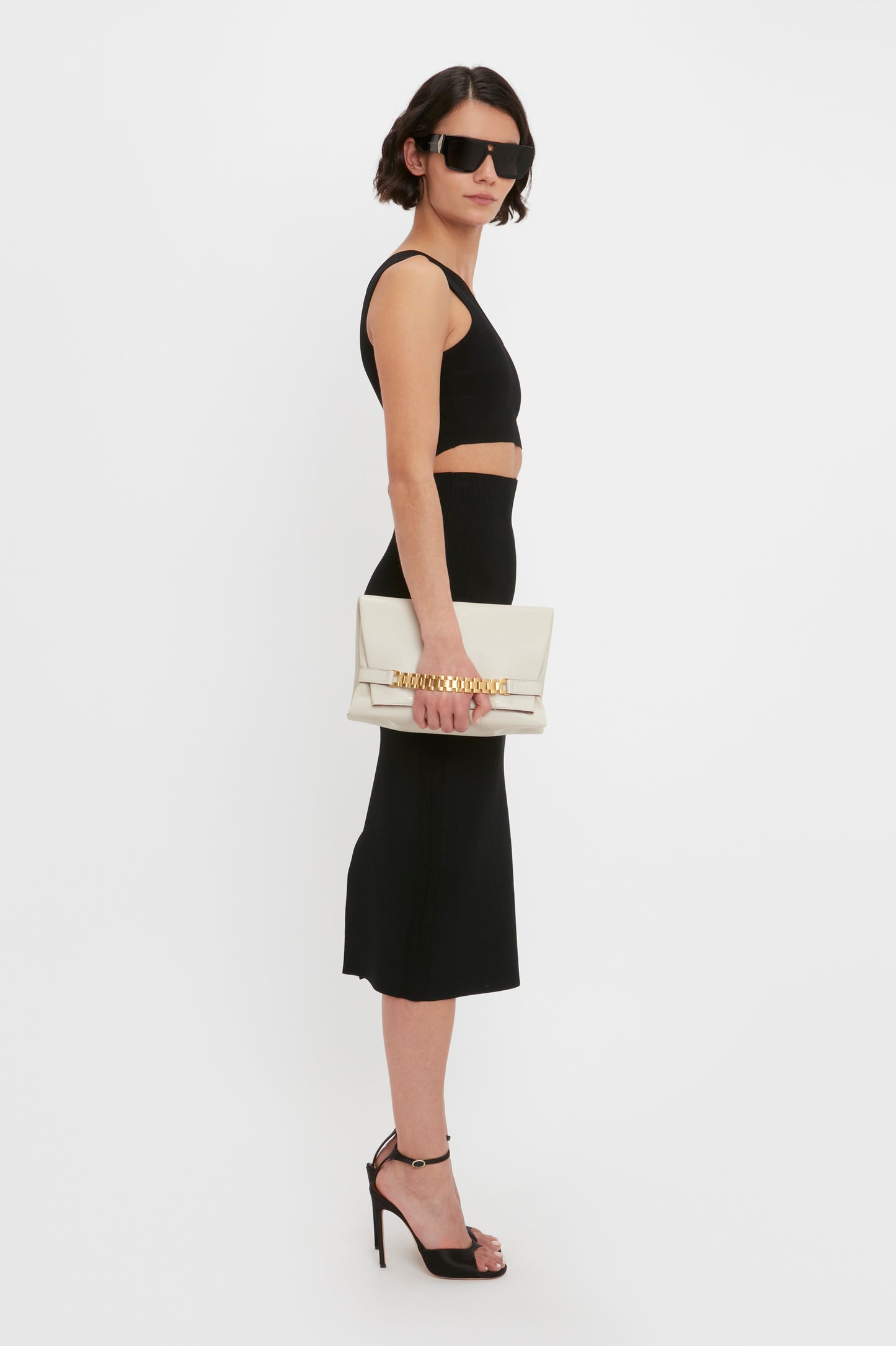 Woman wearing the VB Body One Shoulder Crop Top in Black by Victoria Beckham, paired with a midi skirt, black high heels, and sunglasses. She is holding a beige clutch bag and standing in front of a plain white background.