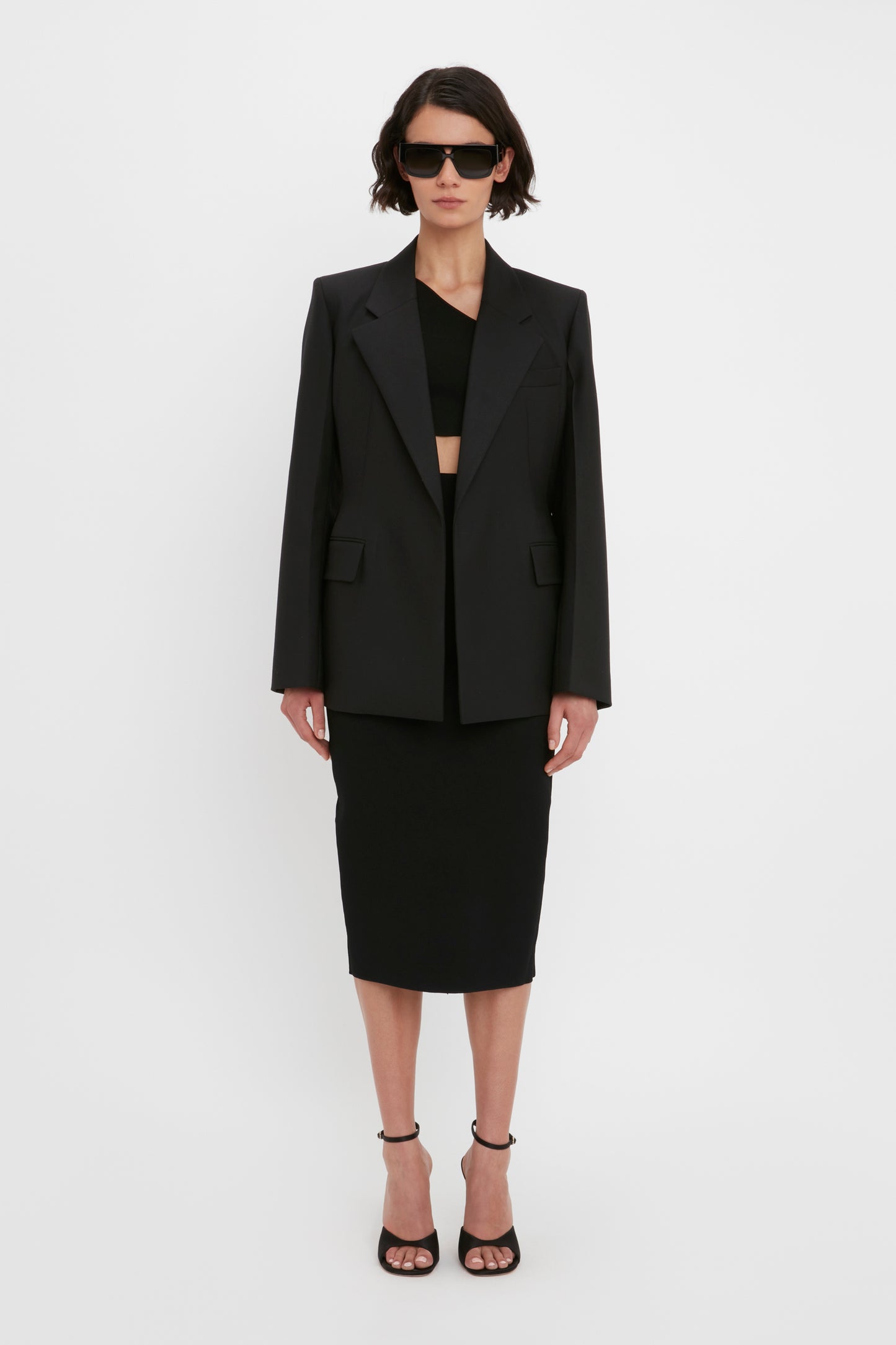 Person wearing a Victoria Beckham One Shoulder Crop Top in Black, paired with a black blazer and black pencil skirt. They have short hair and are wearing black sunglasses and black high-heeled sandals.