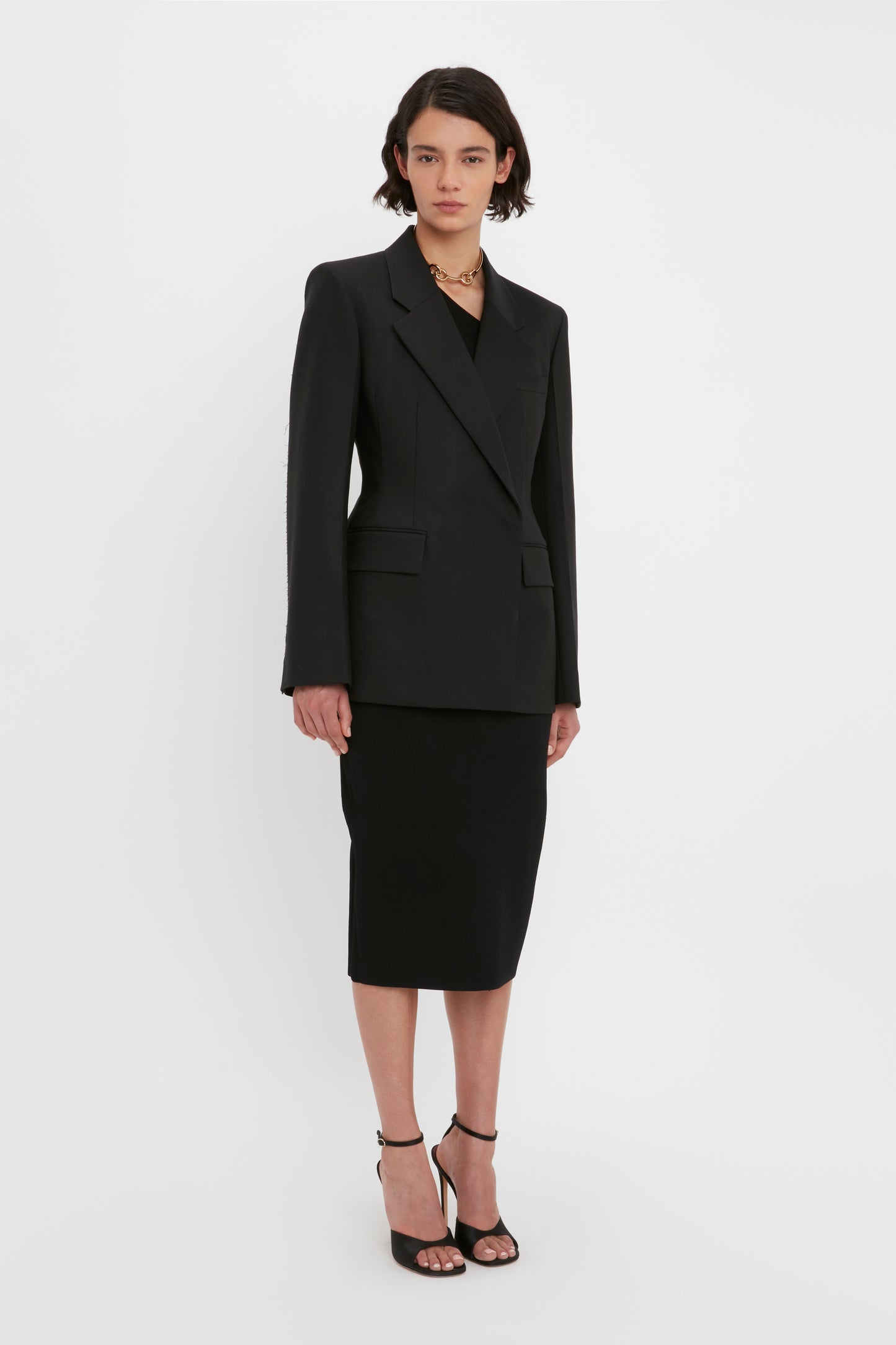 A person with short dark hair wearing a black Victoria Beckham VB Body One Shoulder Crop Top, a black blazer, black skirt, and black high-heeled sandals stands against a plain white background.
