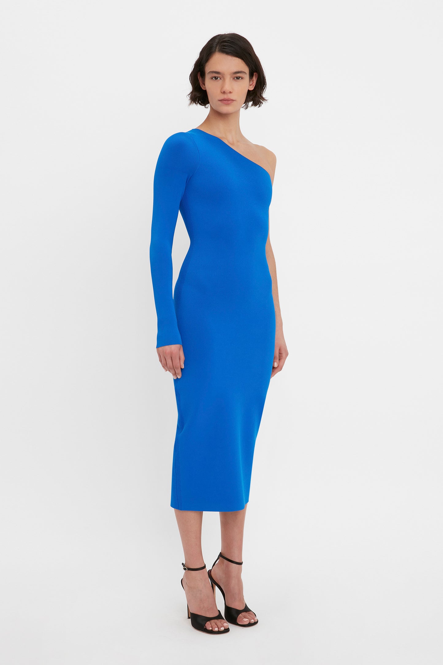 A woman stands wearing the Victoria Beckham VB Body One Shoulder Midi Dress in Cobalt, a body-hugging design featuring a single long sleeve. She pairs the dress with black high-heeled sandals against a plain white background.