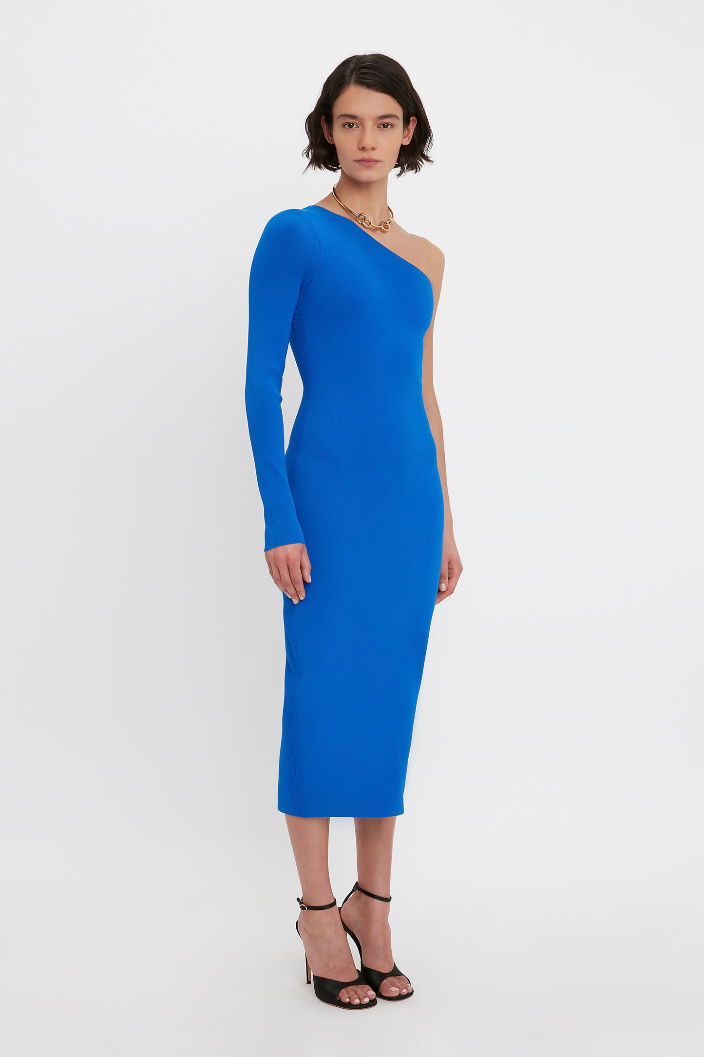 A person stands wearing the VB Body One Shoulder Midi Dress in Cobalt by Victoria Beckham, paired with black high-heeled sandals, with a neutral expression.