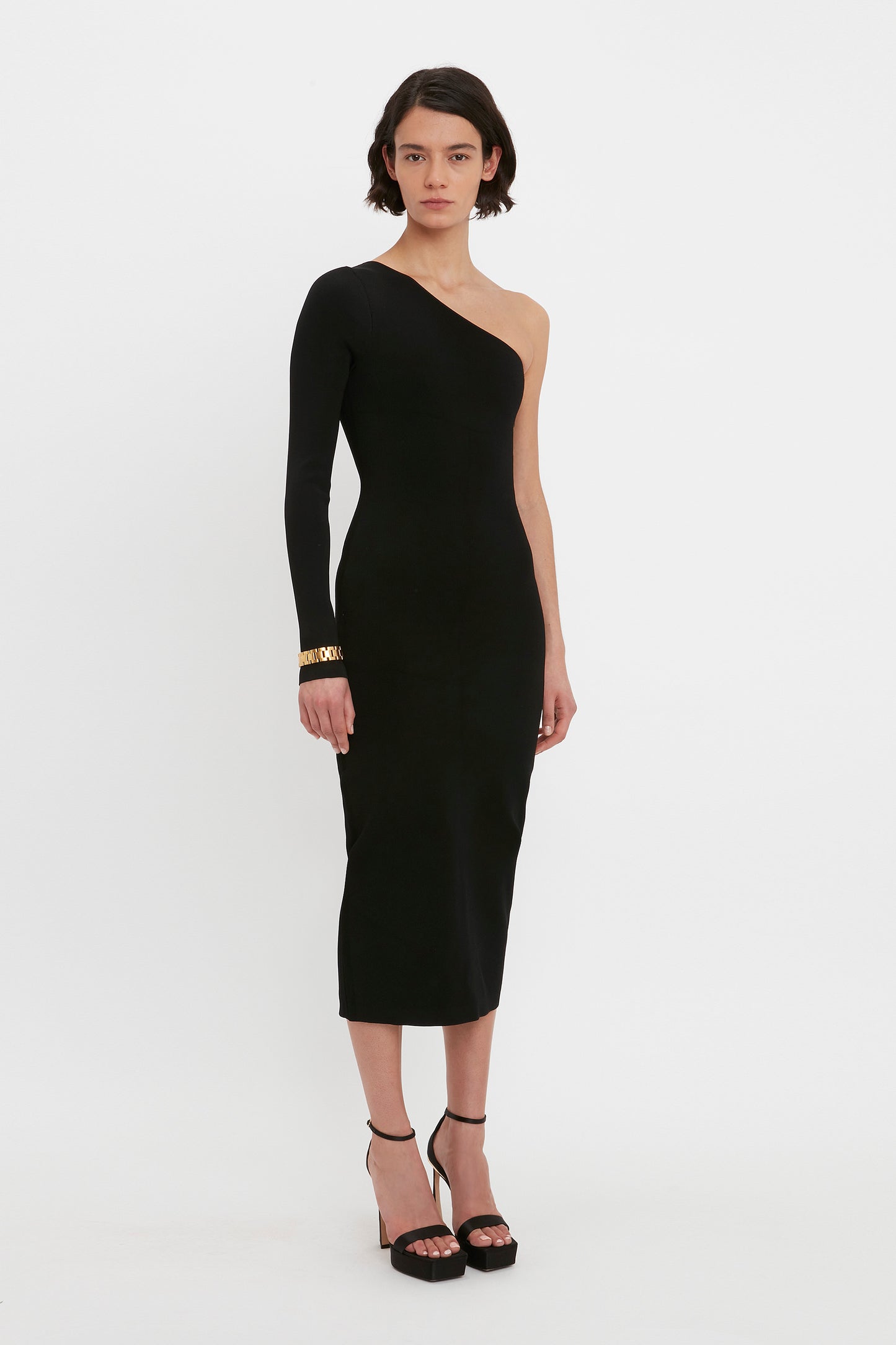A person with short hair stands against a white background wearing the VB Body One Shoulder Midi Dress in Black by Victoria Beckham, paired with black high-heeled shoes.
