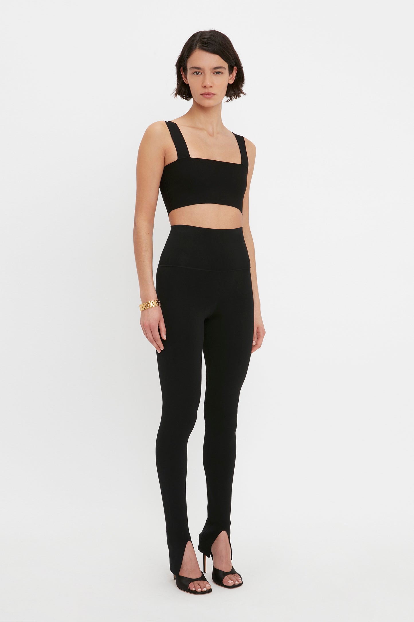 A person stands against a white background, wearing the VB Body Strap Bandeau Top in Black by Victoria Beckham and matching high-waisted leggings with stirrups, along with black heeled sandals and a gold bracelet.