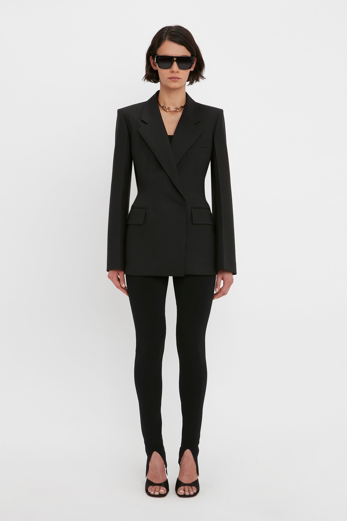 A person in a black blazer, VB Body Split Front Leggings in Black by Victoria Beckham, and black heels stands against a white background, wearing dark sunglasses and a chain necklace.