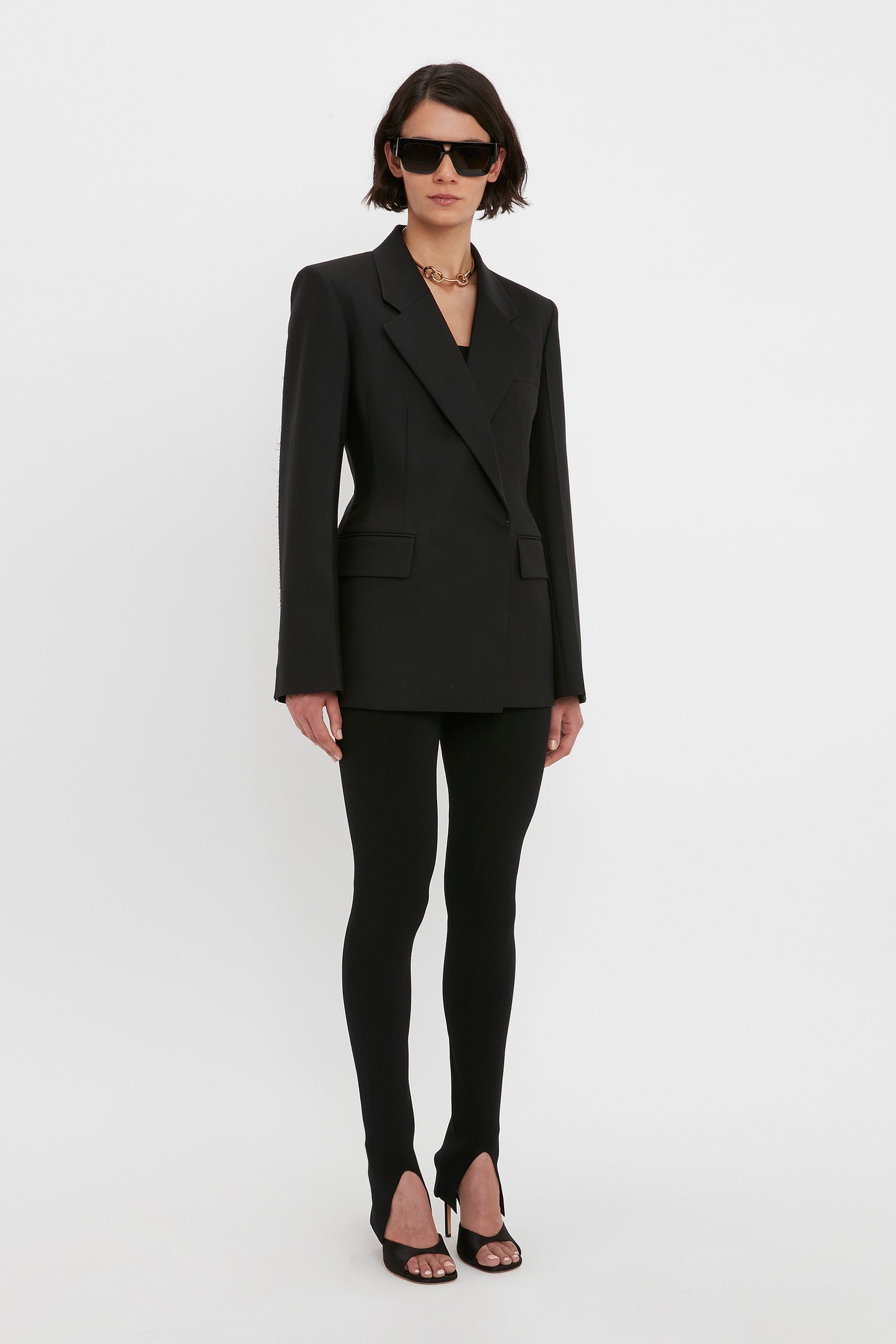 Person wearing a black blazer, VB Body Split Front Leggings in Black by Victoria Beckham, black sandals, sunglasses, and a gold chain necklace, standing against a plain white background.