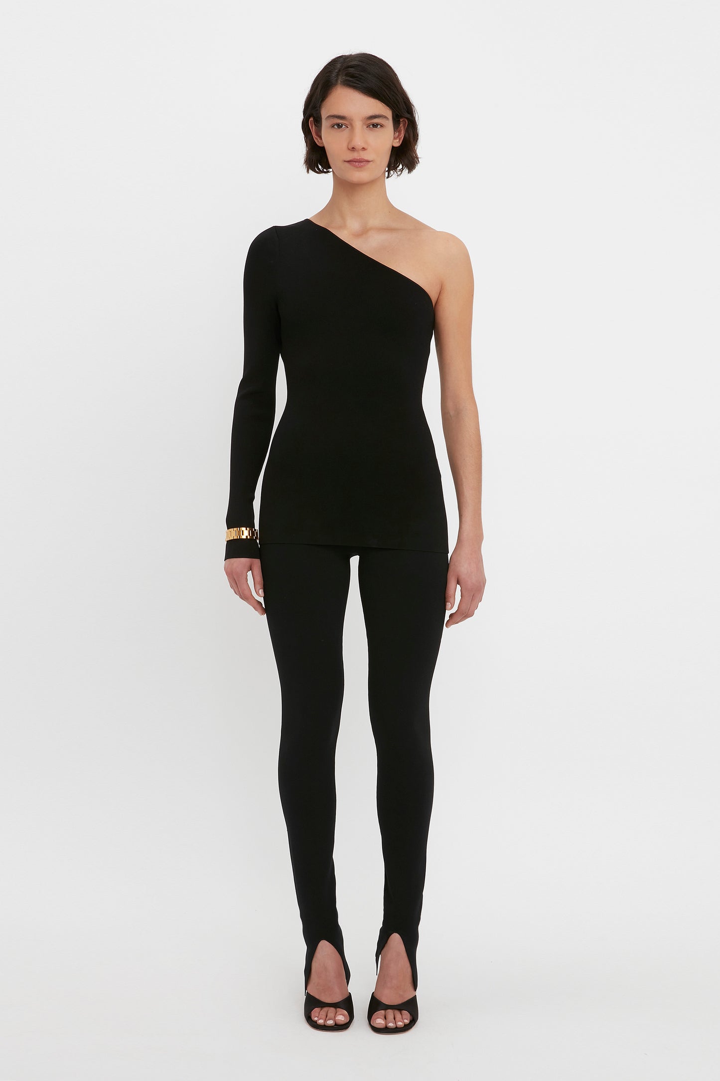 A person stands against a white background wearing the VB Body One Shoulder Top in Black by Victoria Beckham along with matching black, fitted pants. They are also wearing black shoes with open toes.