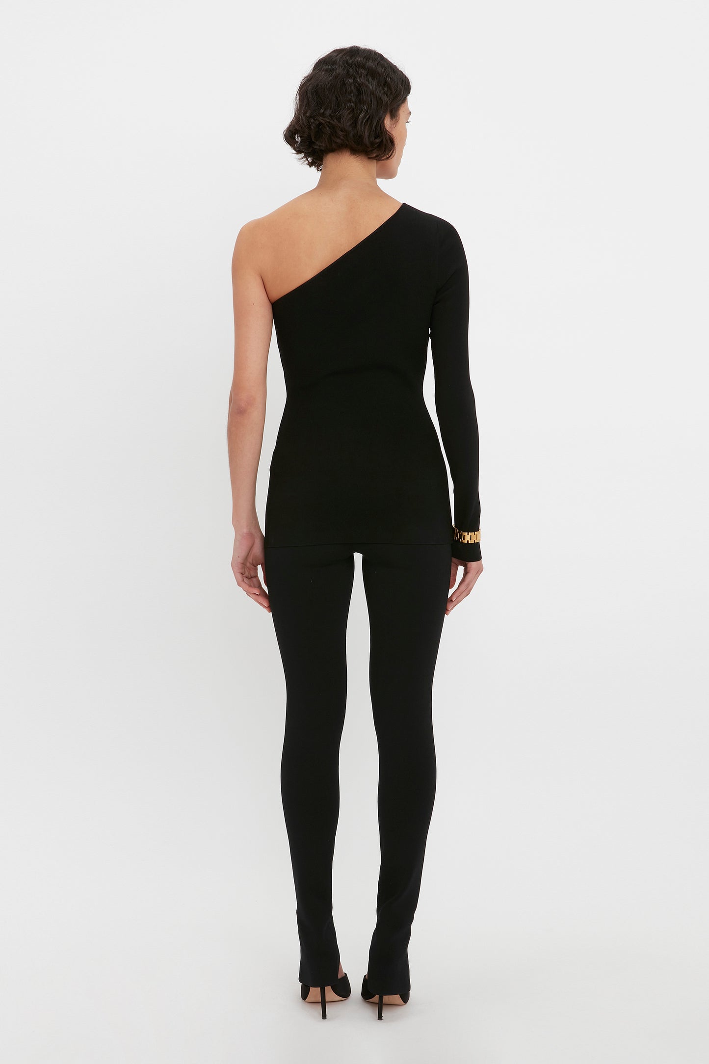 A person stands against a plain background wearing the VB Body One Shoulder Top in Black by Victoria Beckham, showcasing a bare left shoulder and paired with full-length pants, viewed from the back.