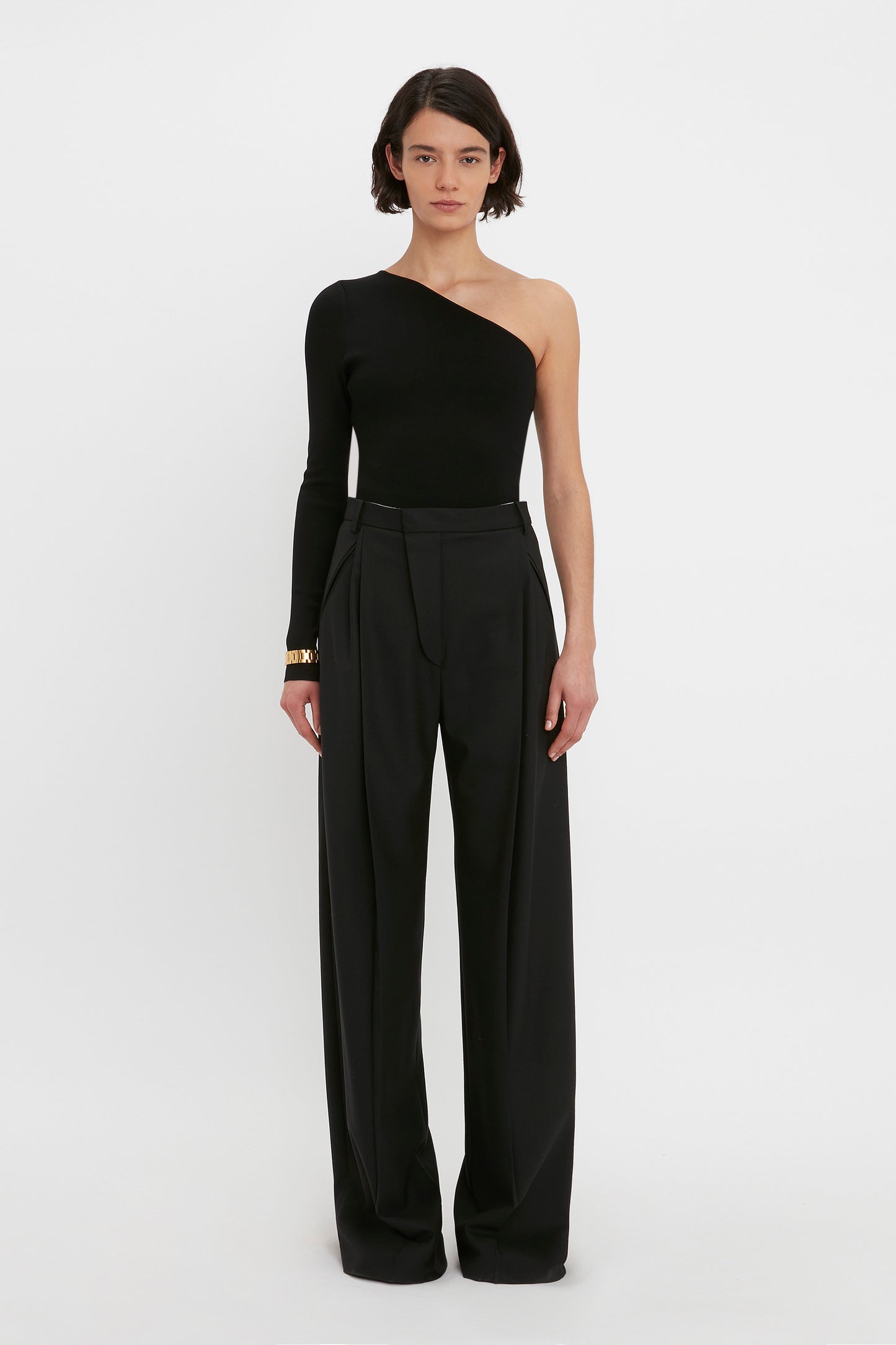 Person standing against a plain white background, wearing the Victoria Beckham VB Body One Shoulder Top in Black and wide-leg black trousers.