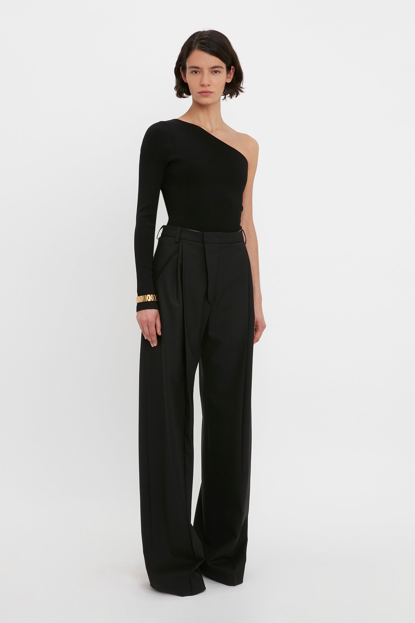Person wearing the VB Body One Shoulder Top in Black by Victoria Beckham, paired with high-waisted, wide-leg black trousers, standing against a plain white background.