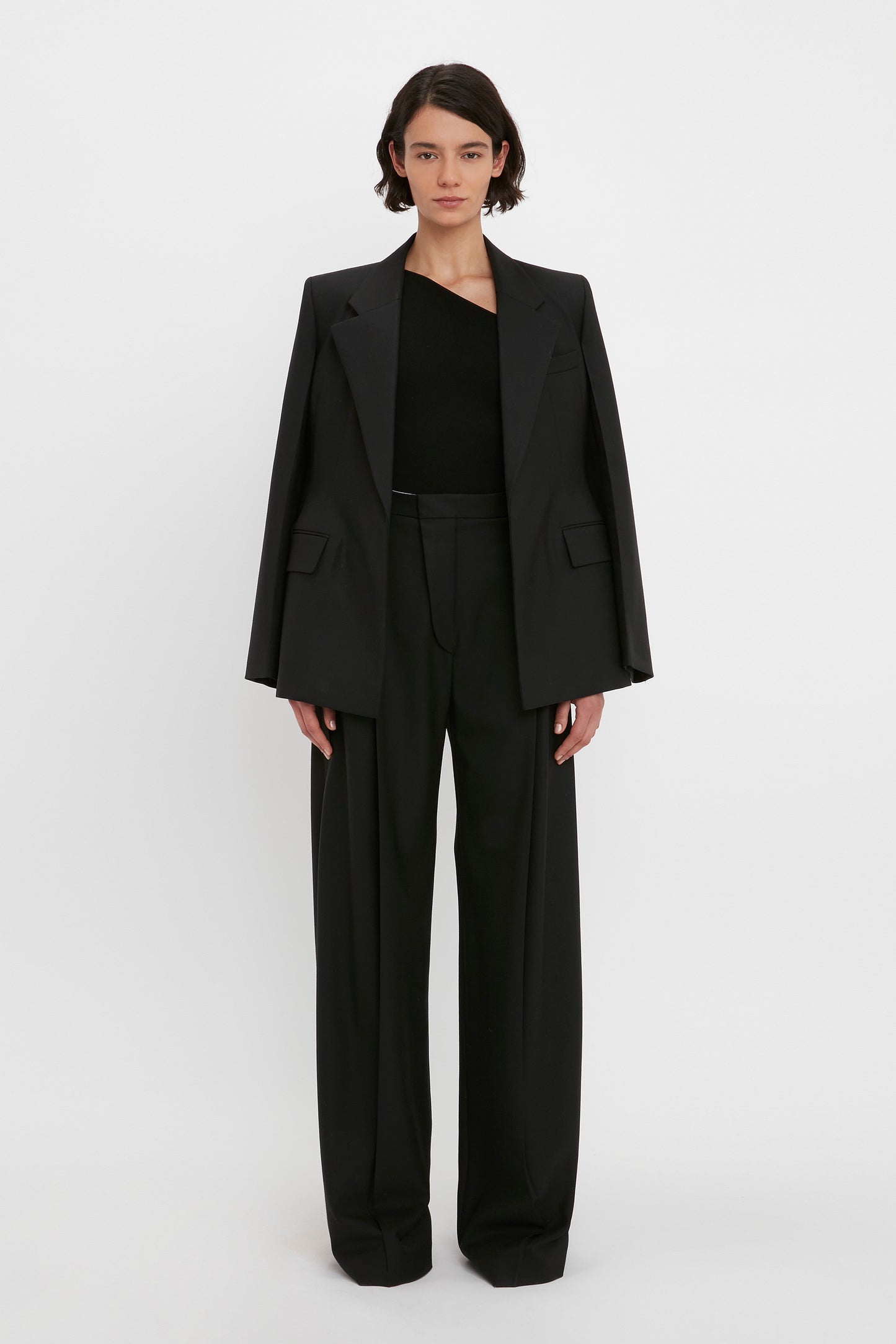A person stands against a white background wearing a black suit jacket draped over their shoulders, paired with black trousers and the VB Body One Shoulder Top in Black by Victoria Beckham.