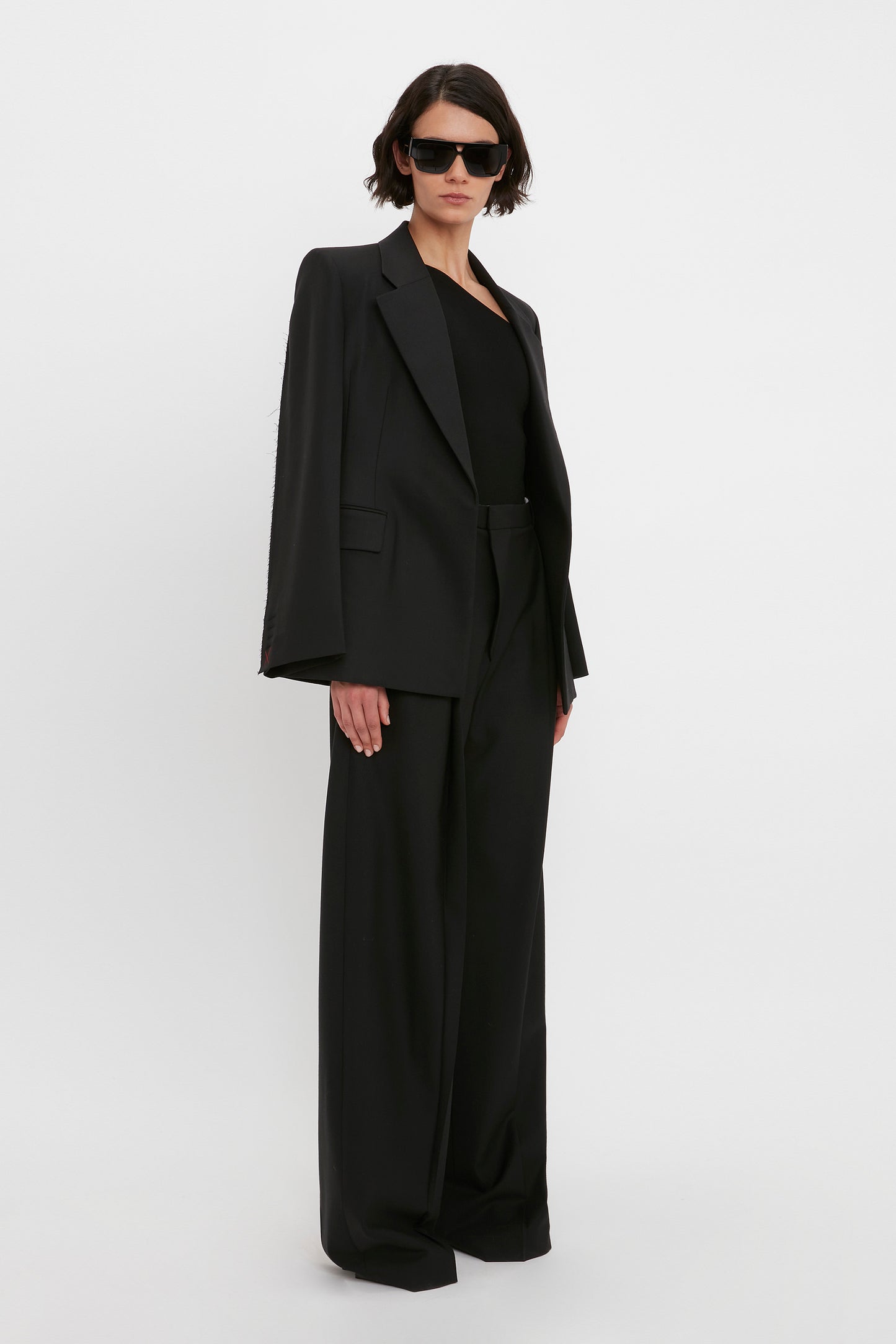 A person wearing a black suit, the VB Body One Shoulder Top in Black by Victoria Beckham, and sunglasses stands against a plain white background.