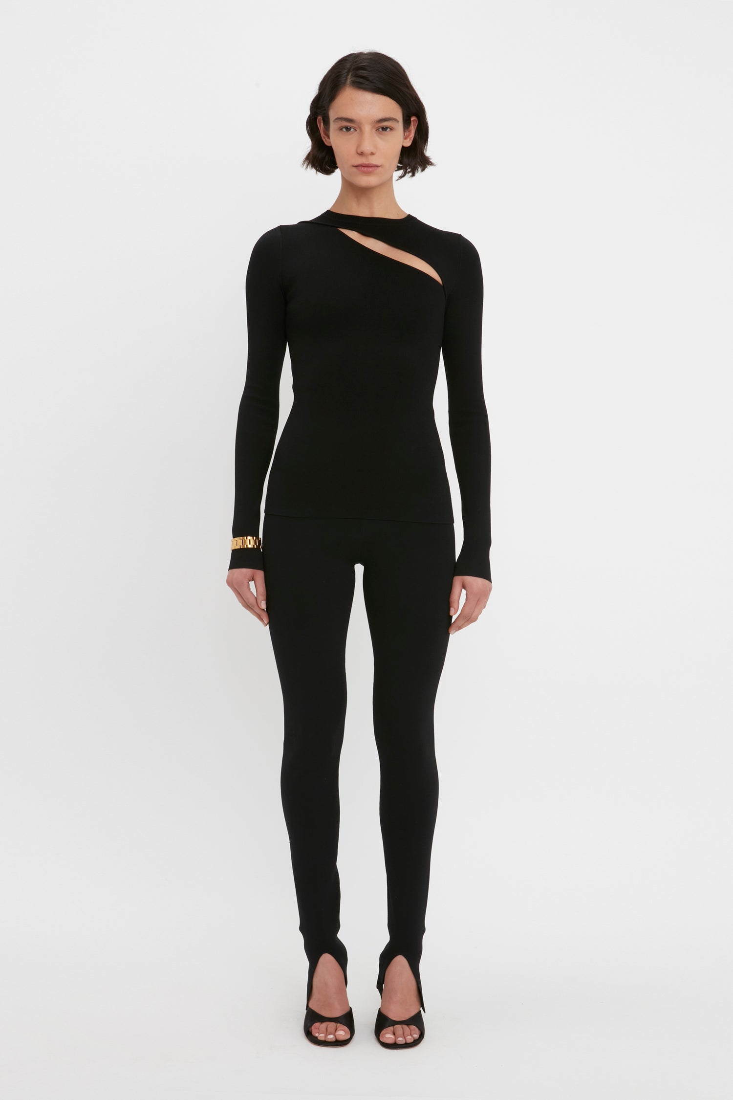 A person stands against a white background wearing the Victoria Beckham VB Body Front Slash Crew Neck Top in Black and matching black pants. They have short, dark hair and are wearing heeled sandals.