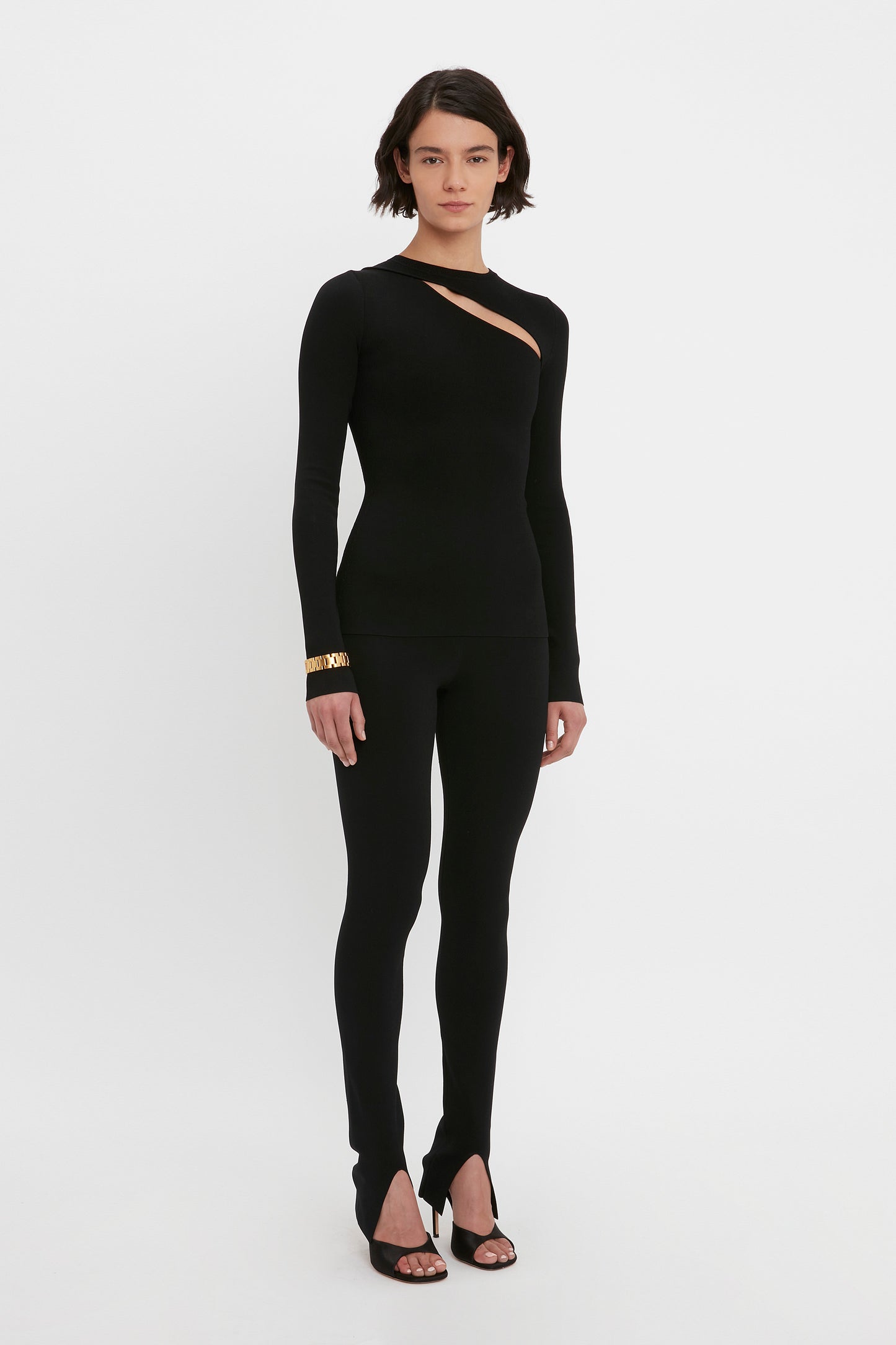 A person stands against a white background wearing a black, long-sleeve VB Body Front Slash Crew Neck Top by Victoria Beckham. The outfit is form-fitting with a diagonal cutout on the chest. They have short, dark hair, and are accessorized with black heels and a gold bracelet.