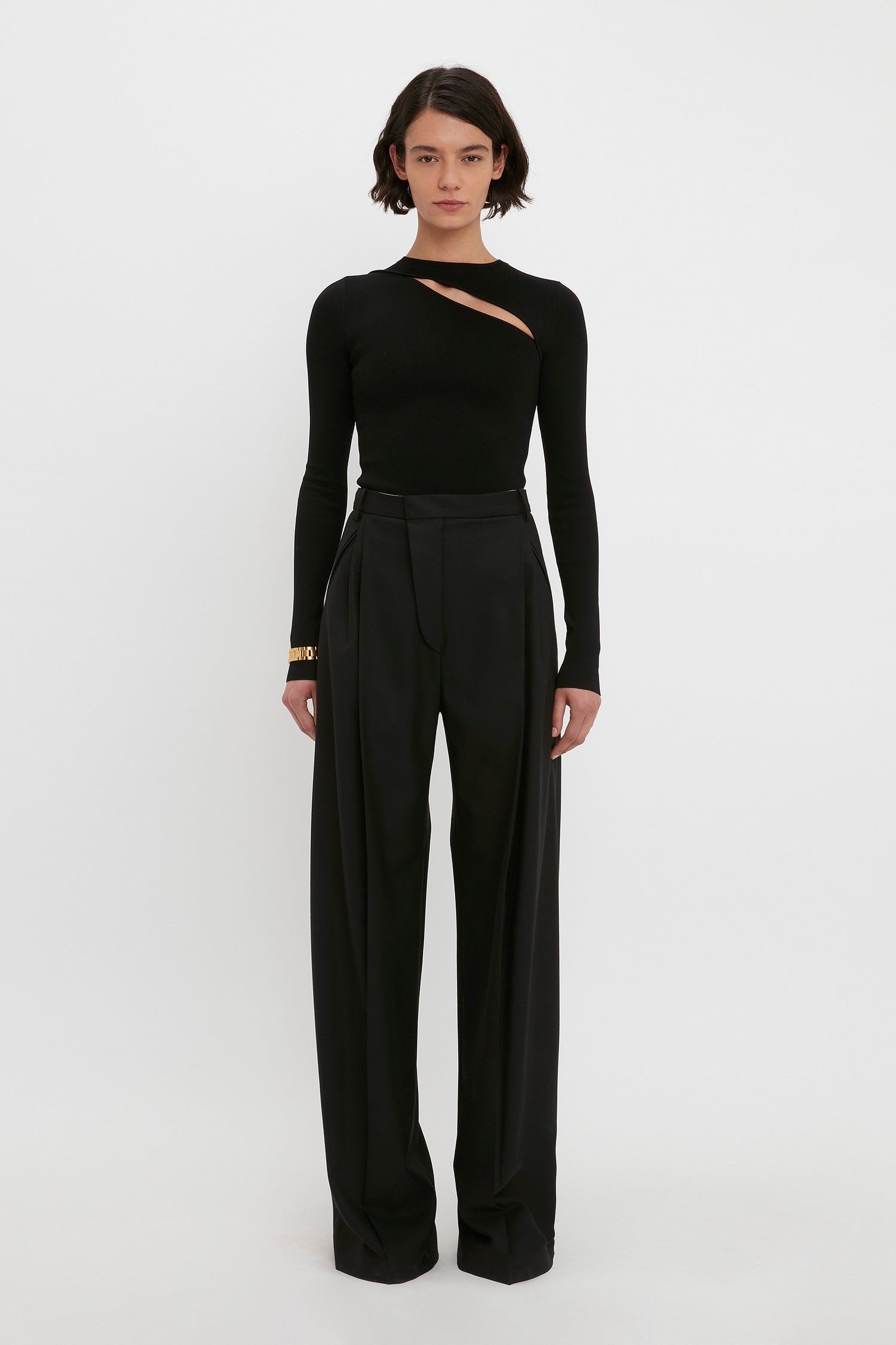 A person stands against a white background wearing the VB Body Front Slash Crew Neck Top in Black from Victoria Beckham and high-waisted black trousers.