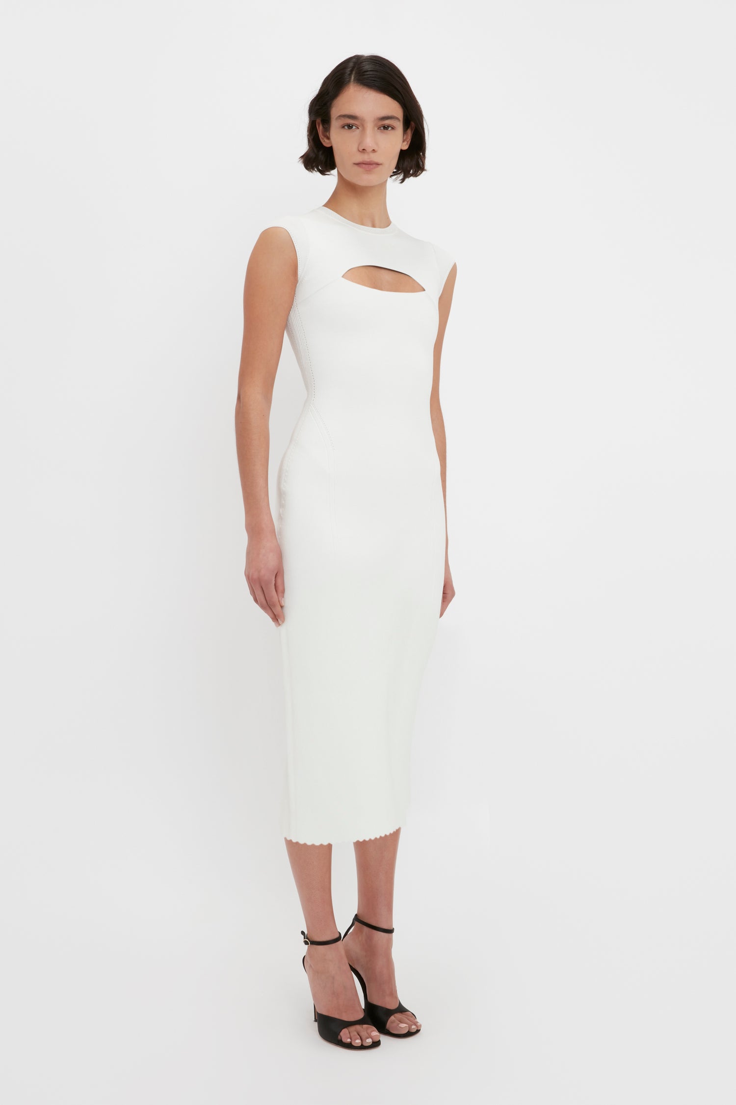 A person stands against a white background wearing the Victoria Beckham VB Body Slash Front Midi Dress in White, paired with black high-heeled sandals.
