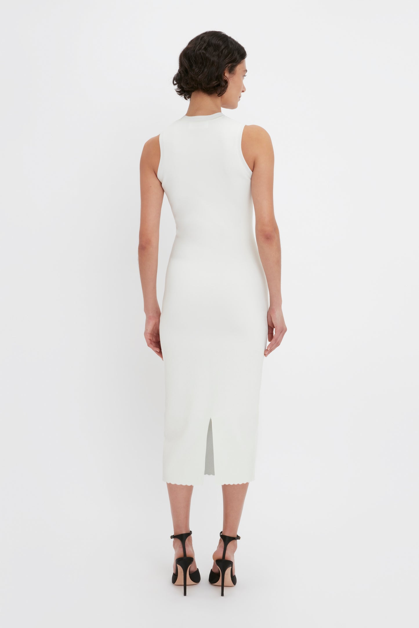 A person with short dark hair is seen from the back, wearing a sleeveless, form-fitting VB Body Slash Front Midi Dress in White by Victoria Beckham and black high heels.
