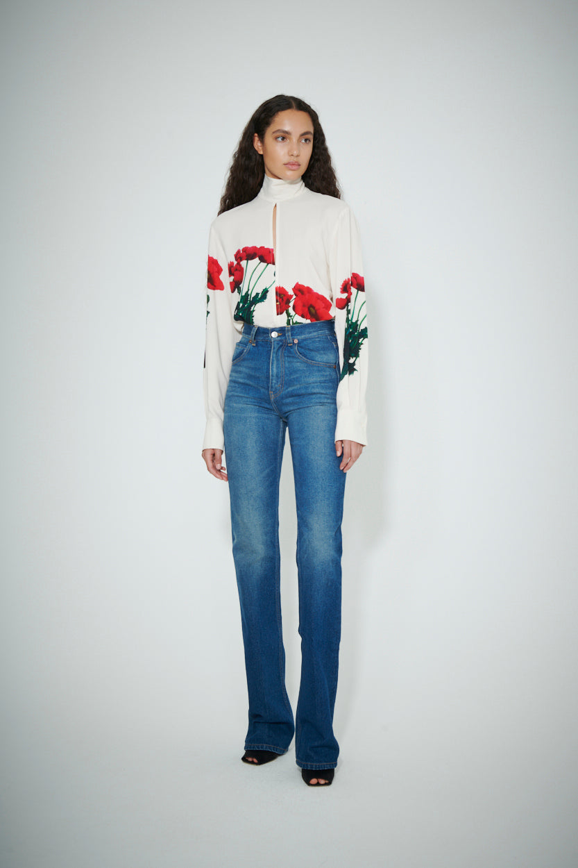A person stands against a plain backdrop wearing the MAW22 3 white turtleneck with a red floral pattern by Victoria Beckham and high-waisted blue jeans.