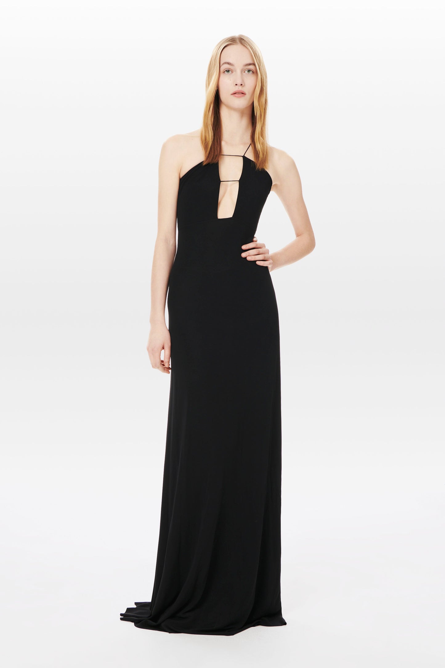 A woman stands wearing a Halter Floor-Length Dress in Black by Victoria Beckham, made from sustainable viscose yarns, featuring a halter neckline and a front cutout, against a plain white background.