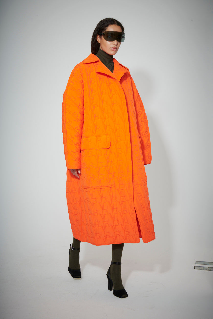 Person wearing a MAW22 2 coat by Victoria Beckham, black turtleneck, black leggings, and black high-heeled boots, with large dark sunglasses, standing against a plain white background.