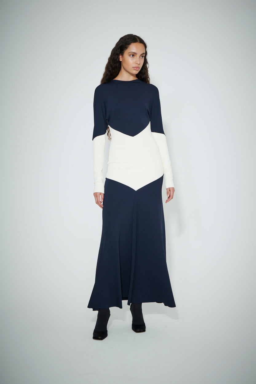 A woman with long curly hair is modeling the Victoria Beckham MAW22 1, a floor-length, long-sleeve navy dress adorned with white geometric accents, paired with black ankle boots. She is standing against a plain light-colored backdrop.