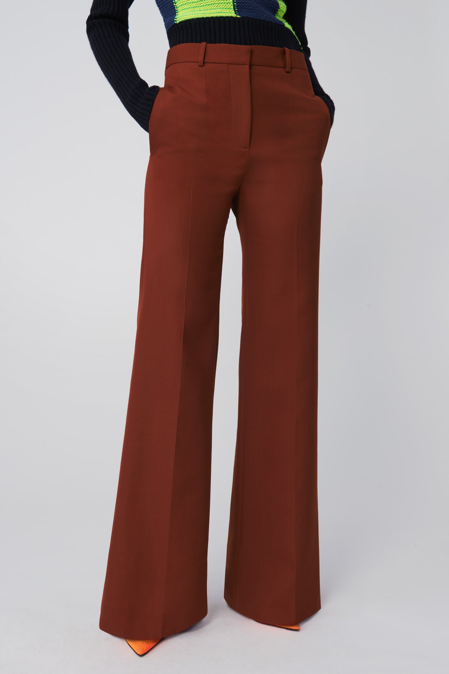 A person models Victoria Beckham's High Waisted Wide Leg Trousers in Chestnut, with hands in pockets against a neutral background.