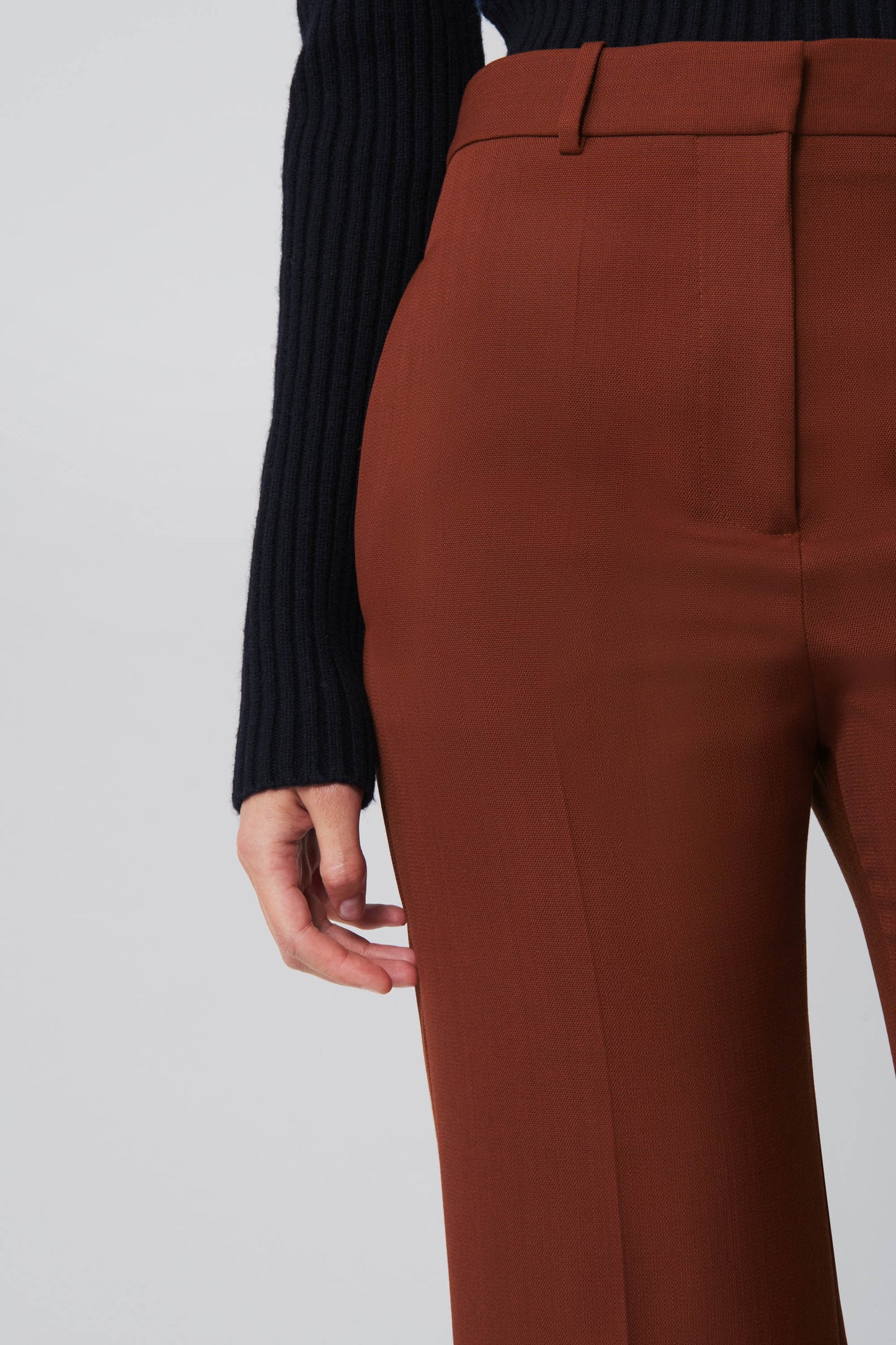 A person is wearing a black ribbed sweater and Victoria Beckham's High Waisted Wide Leg Trousers in Chestnut. The photo emphasizes the lower torso and right hand, showcasing the wool-blend fabric and pants' wide-leg style.