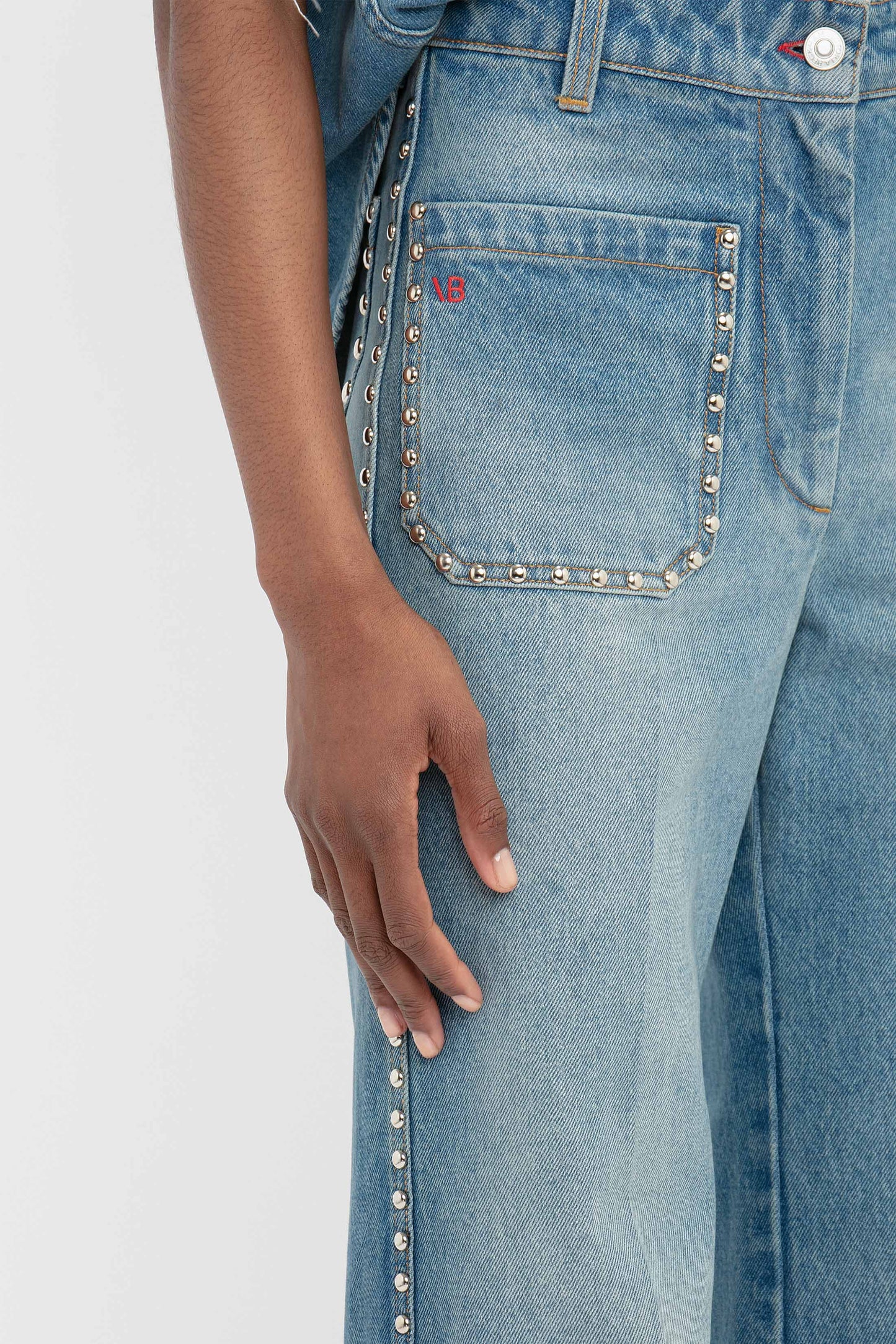 A hand grazes the side of blue denim, highlighting the high waist and elegant studded details along the pocket and seam of Victoria Beckham's BAO TEST R - Alina High Waisted Jean In Vintage Wash, exuding a vintage charm.