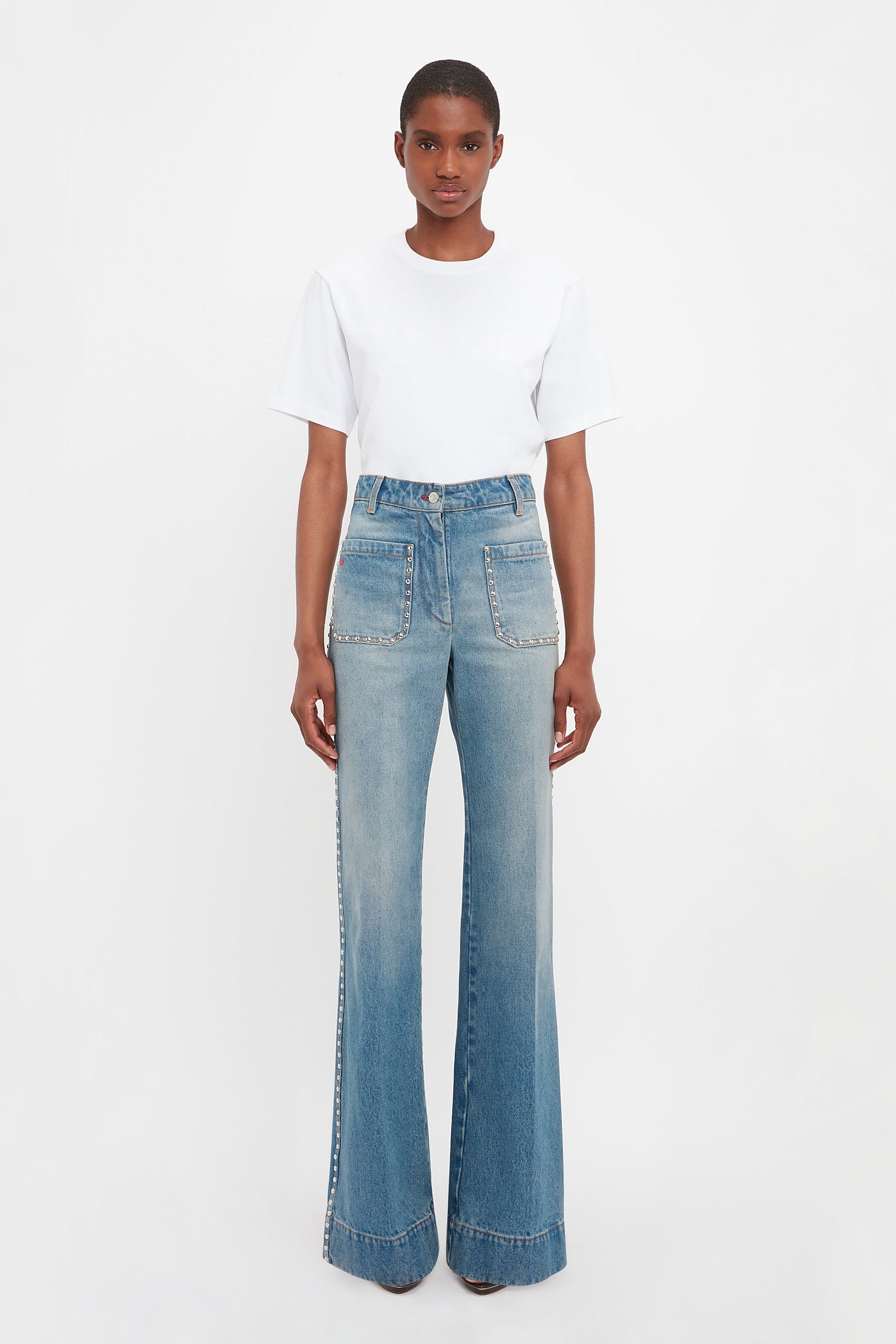 Someone stands facing forward, wearing a white t-shirt and Victoria Beckham's BAO TEST R - Alina High Waisted Jeans in Vintage Wash with front patch pockets.