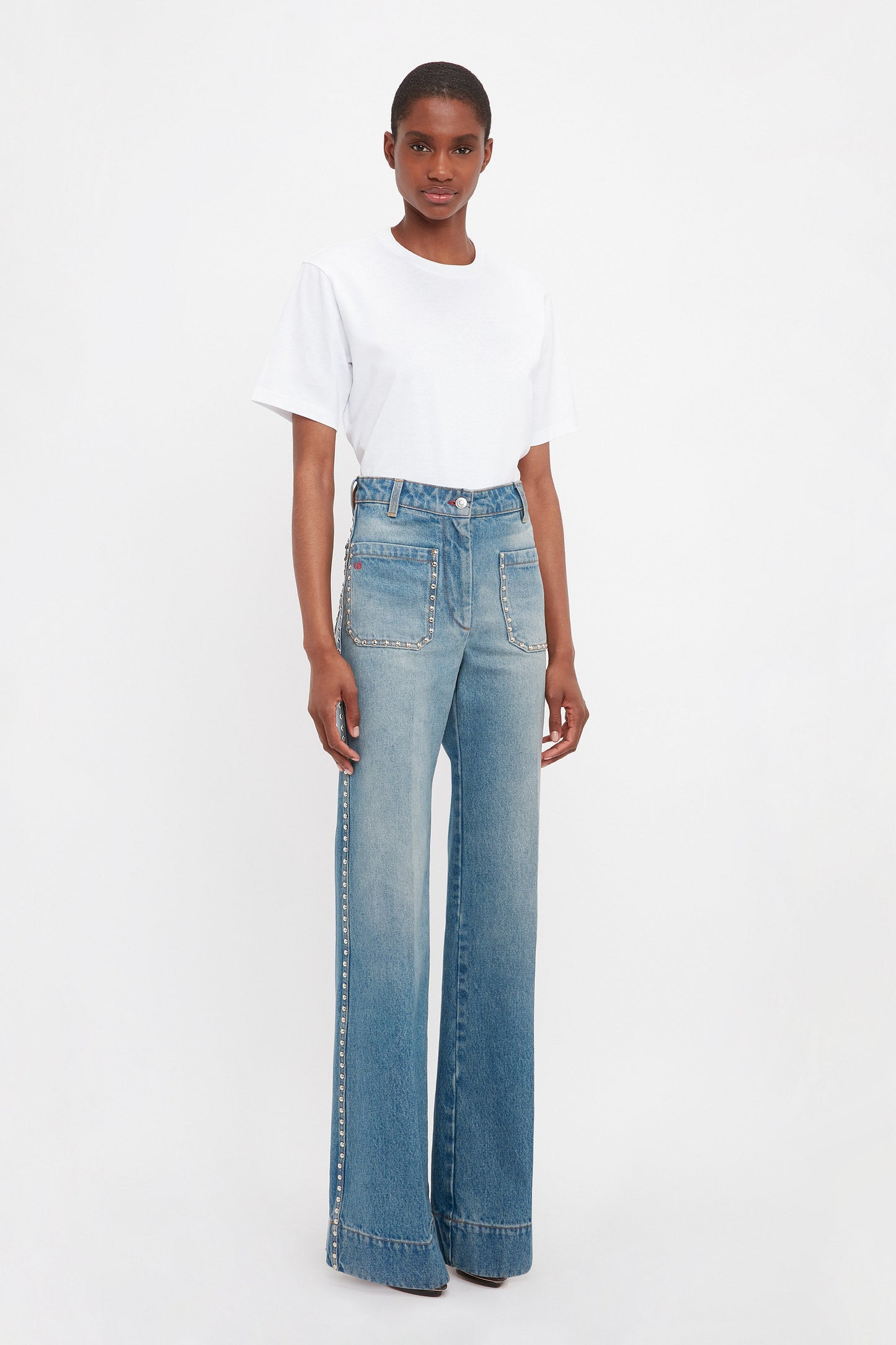 A person in a white T-shirt and Victoria Beckham's BAO TEST R - Alina High Waisted Jean in Vintage Wash, featuring flared legs and side embellishments, stands against a plain white background.