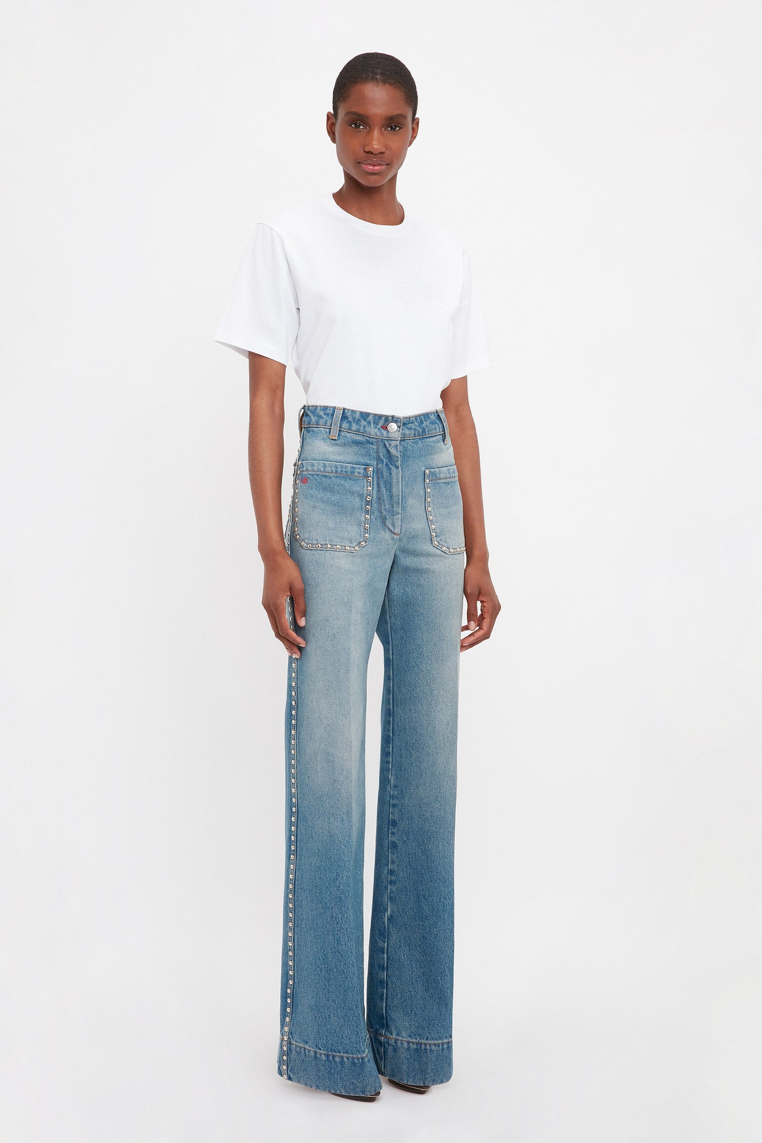 A person in a white T-shirt and Victoria Beckham's BAO TEST R - Alina High Waisted Jean in Vintage Wash, featuring flared legs and side embellishments, stands against a plain white background.
