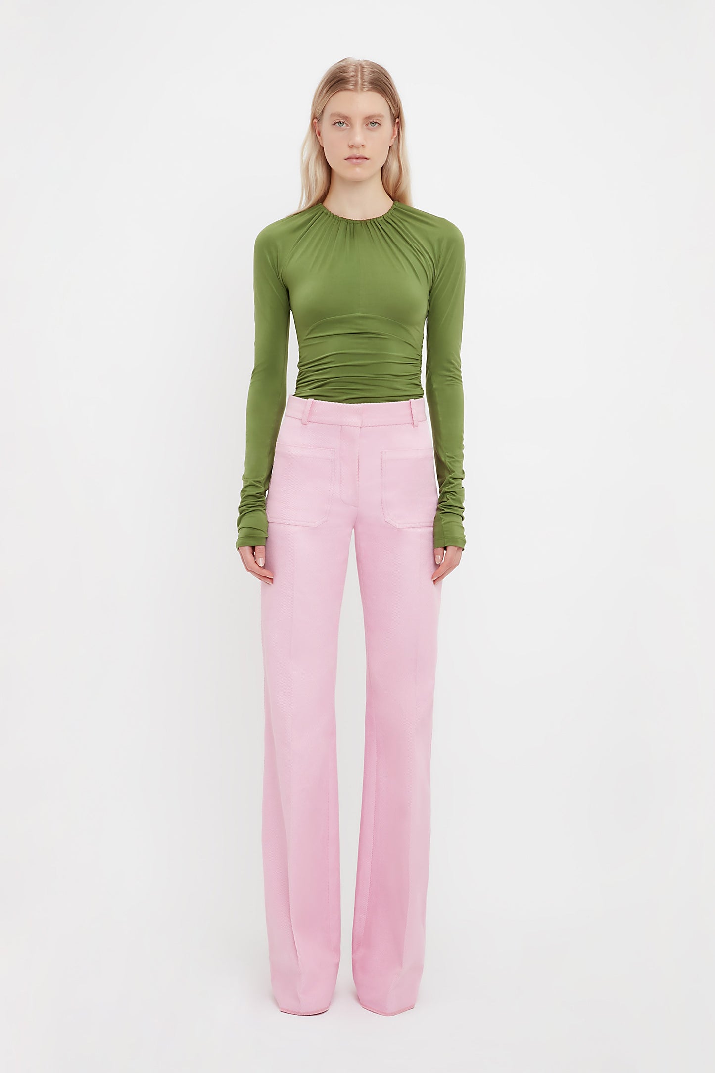 A woman faces forward in a long-sleeve green ruched top and Victoria Beckham’s Alina Tailored Trouser in Bubblegum, featuring a wide-leg 70s silhouette, set against a plain white background.