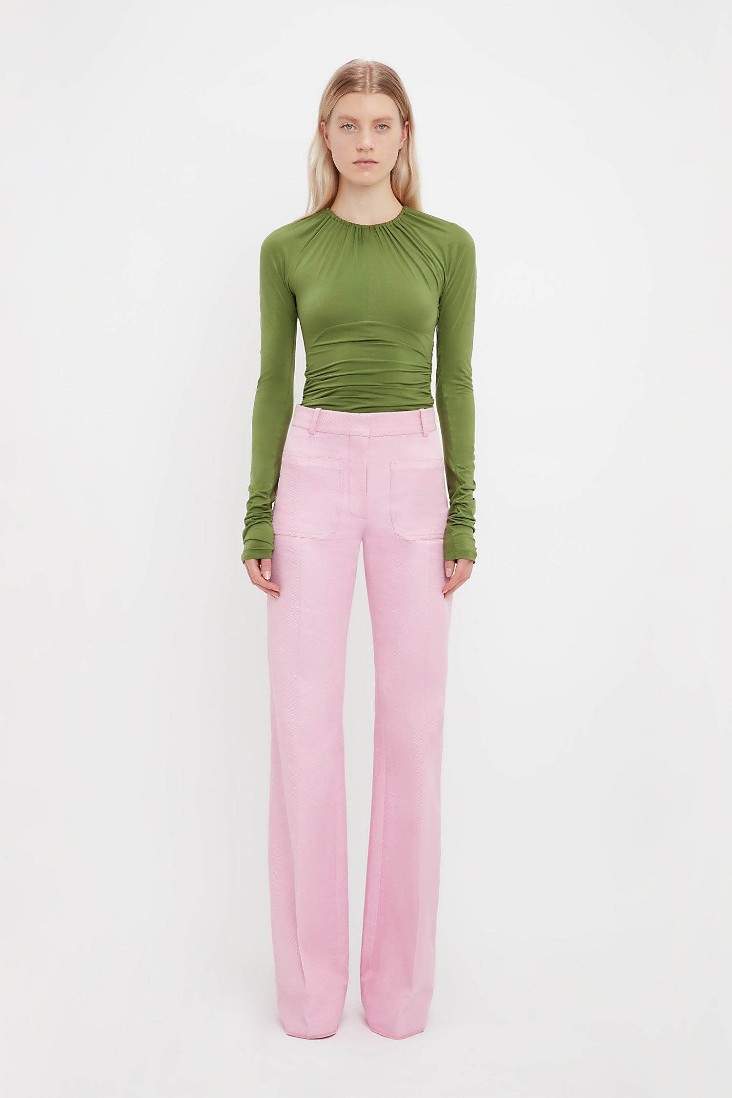 A woman faces forward in a long-sleeve green ruched top and Victoria Beckham’s Alina Tailored Trouser in Bubblegum, featuring a wide-leg 70s silhouette, set against a plain white background.