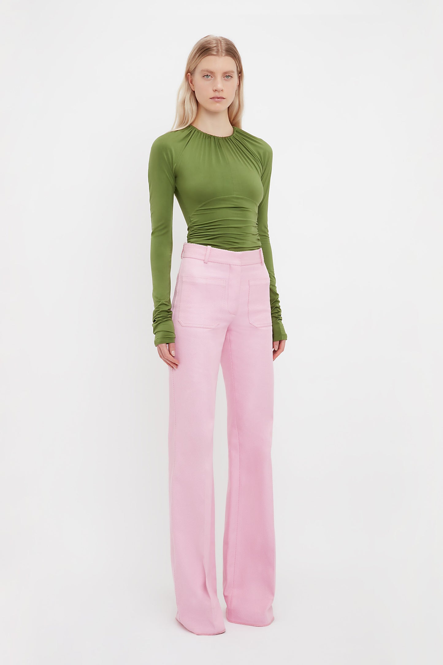 A person poses against a white background in a fitted green long-sleeve top and Victoria Beckham’s Alina Tailored Trouser in Bubblegum, highlighting a chic 70s-inspired silhouette.