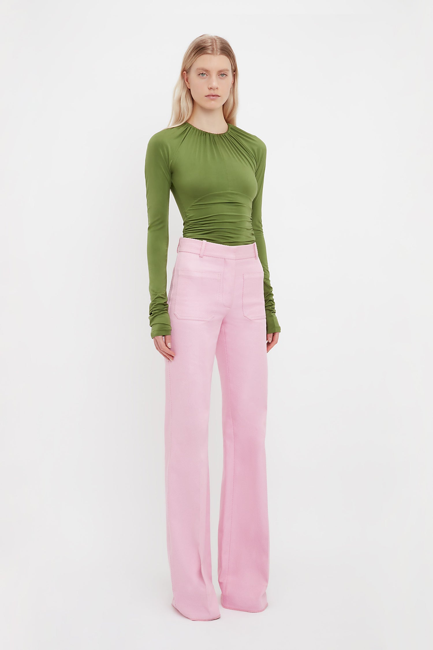 A person poses against a white background in a fitted green long-sleeve top and Victoria Beckham’s Alina Tailored Trouser in Bubblegum, highlighting a chic 70s-inspired silhouette.