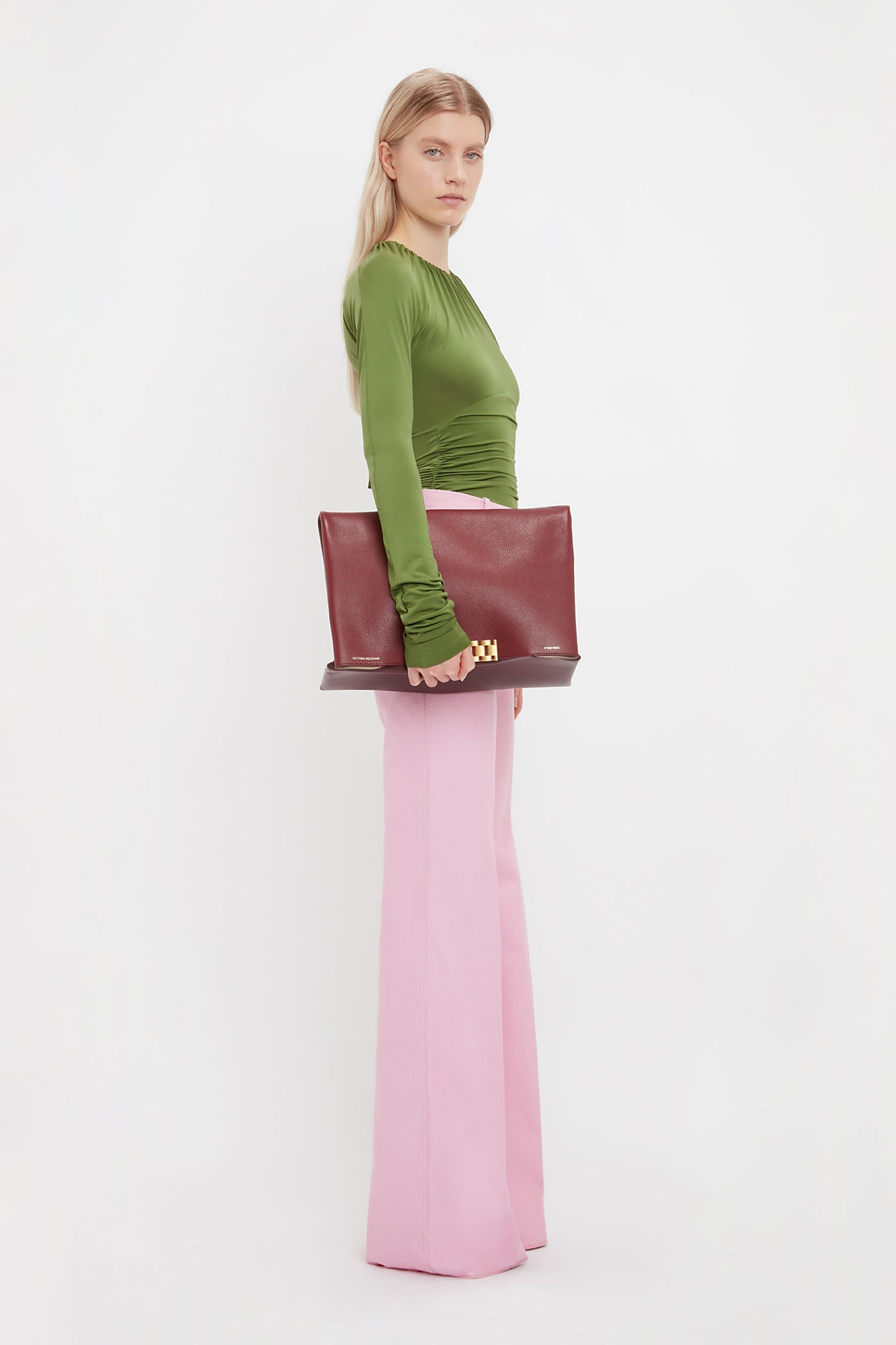 A woman stands against a white background, wearing a green long-sleeve top and Victoria Beckham’s Alina Tailored Trouser in Bubblegum pink, styled with a large burgundy clutch.