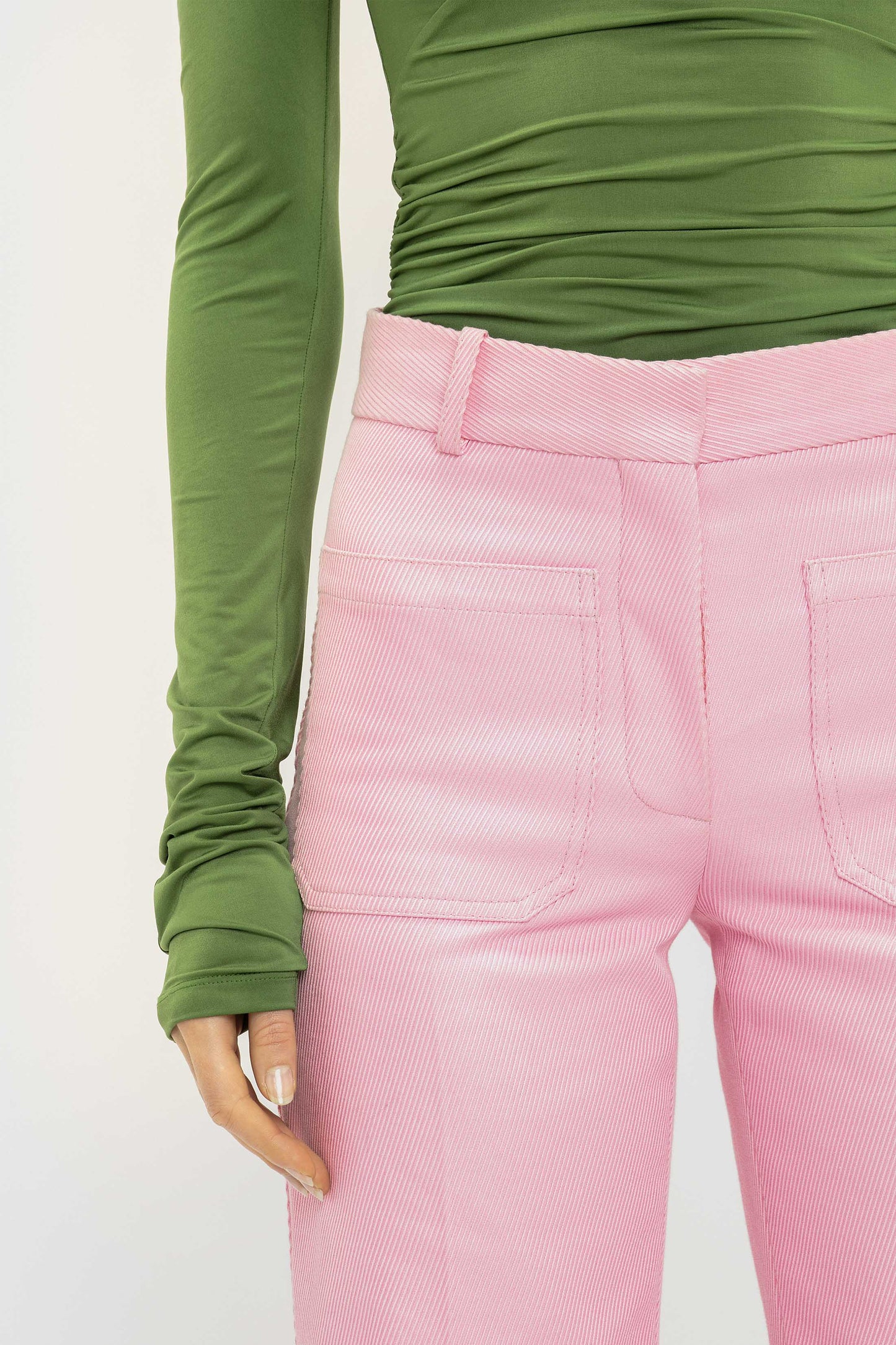 A person wears a long-sleeved green top and Victoria Beckham Alina Tailored Trouser in Bubblegum with large front pockets, creating a chic 70s silhouette. Only the torso and upper legs are visible.