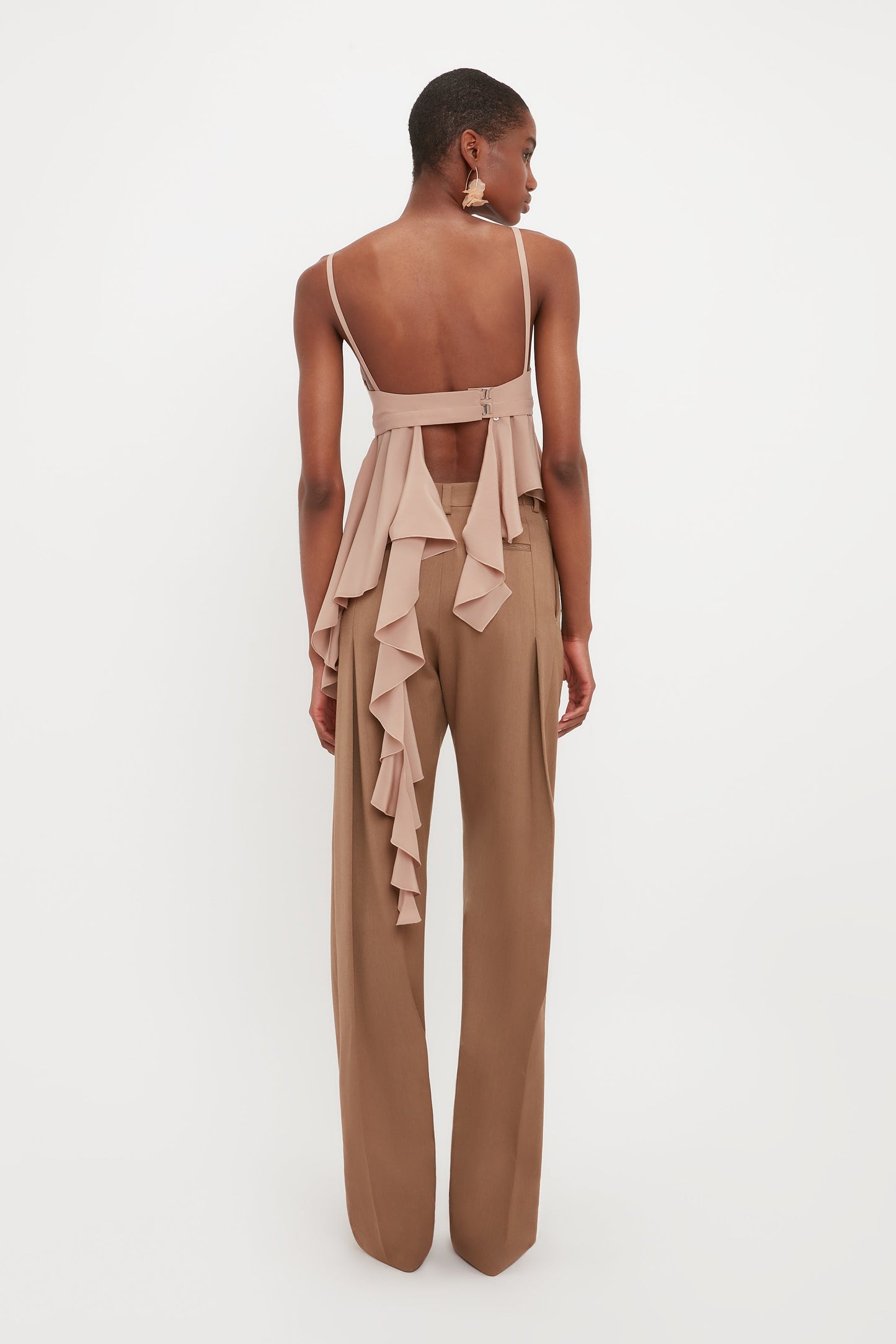 A person stands facing away, wearing a beige ruffled backless top paired with Victoria Beckham’s Front Pleat Trousers in Fawn—an essential wardrobe staple.
