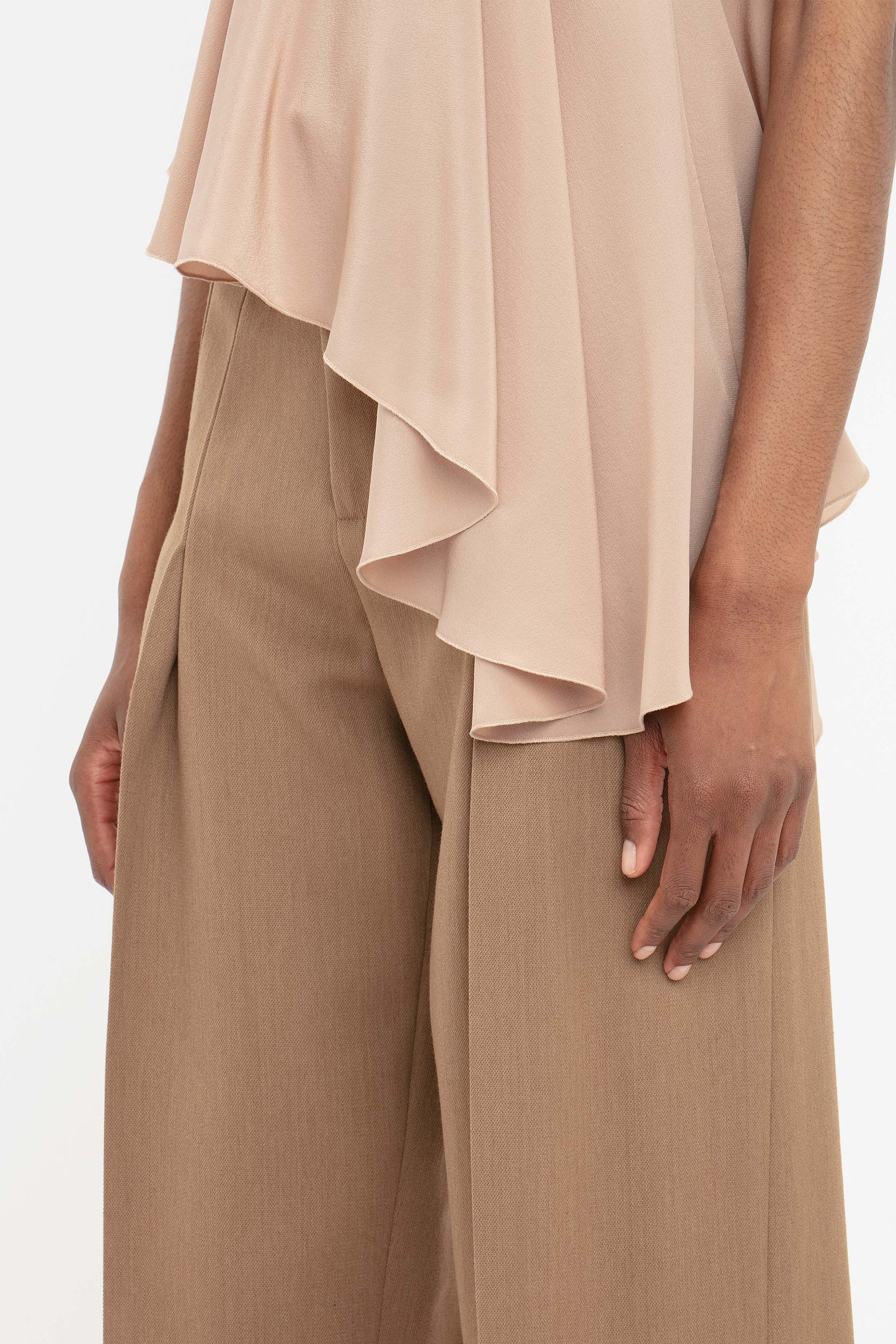 A person wears Victoria Beckham’s Front Pleat Trousers in Fawn—an essential wide-leg pant—paired with a beige asymmetrical top, shown waist down with one hand at their side.
