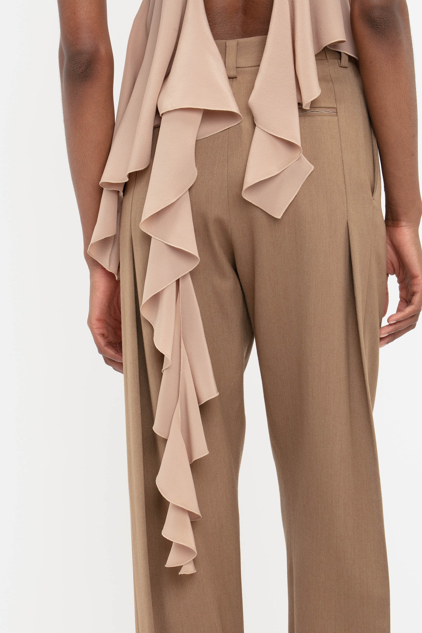 A person shown from behind wears Victoria Beckham's Front Pleat Trousers in Fawn and a sleeveless top with ruffled beige fabric, set against a white background—a true wardrobe essential.