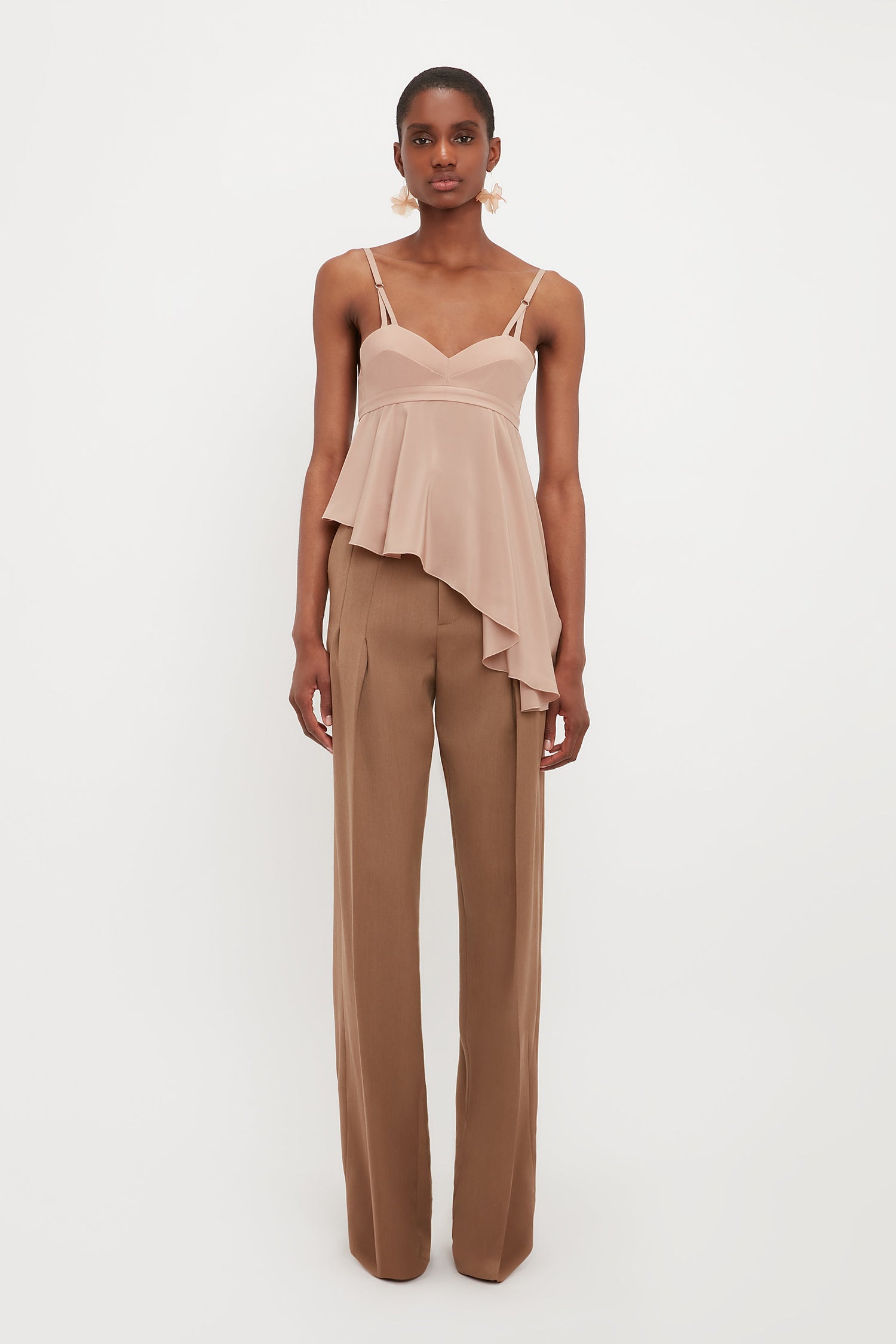 A person stands against a white background in Victoria Beckham’s Front Pleat Trousers in Fawn paired with a blush pink asymmetrical peplum top—an essential wardrobe combination.