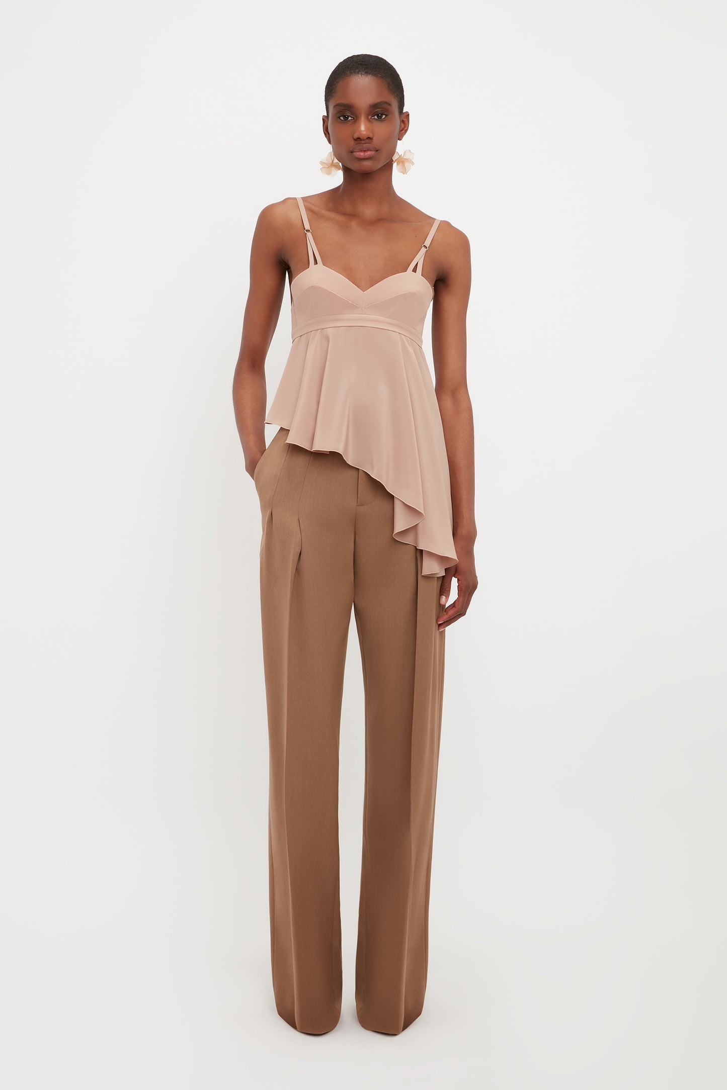 A person stands against a white background wearing a blush asymmetrical camisole top and Victoria Beckham's Front Pleat Trousers In Fawn, styled high-waisted with pleats and wide legs, hands in pockets.