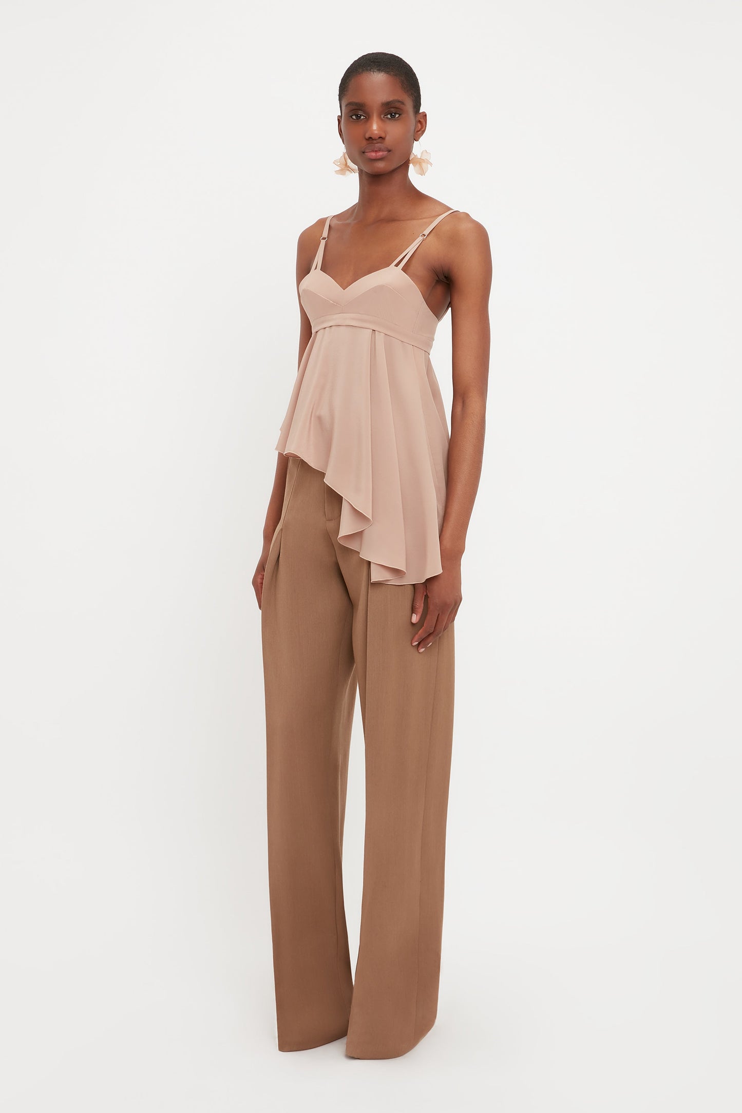 A woman stands against a white background wearing Victoria Beckham’s Front Pleat Trousers In Fawn, styled with a sleeveless, asymmetrical light beige top—a true wardrobe essential.
