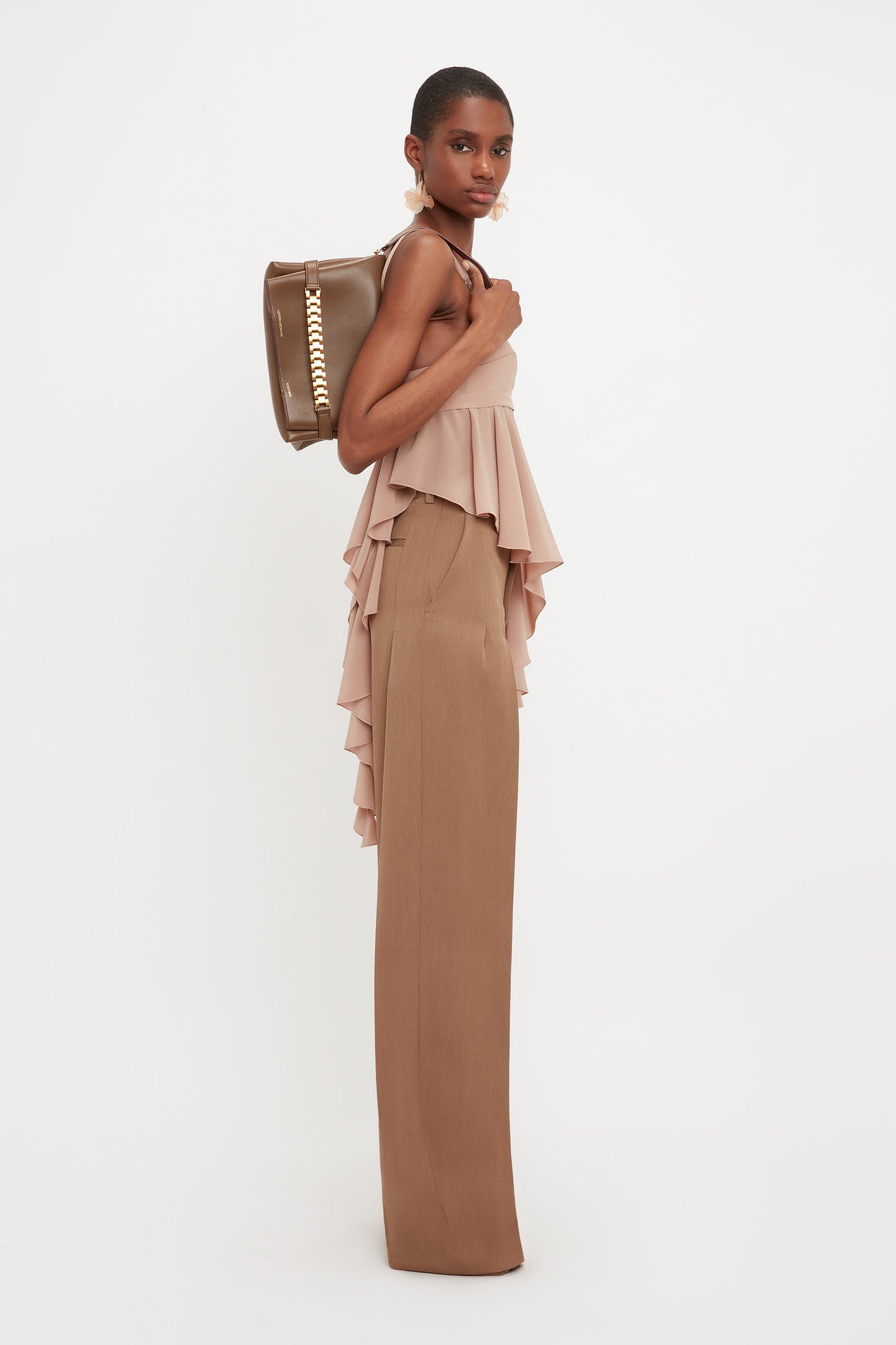 A woman stands in profile wearing a ruffled beige top, Victoria Beckham Front Pleat Trousers in Fawn, and carries a brown handbag over her shoulder.