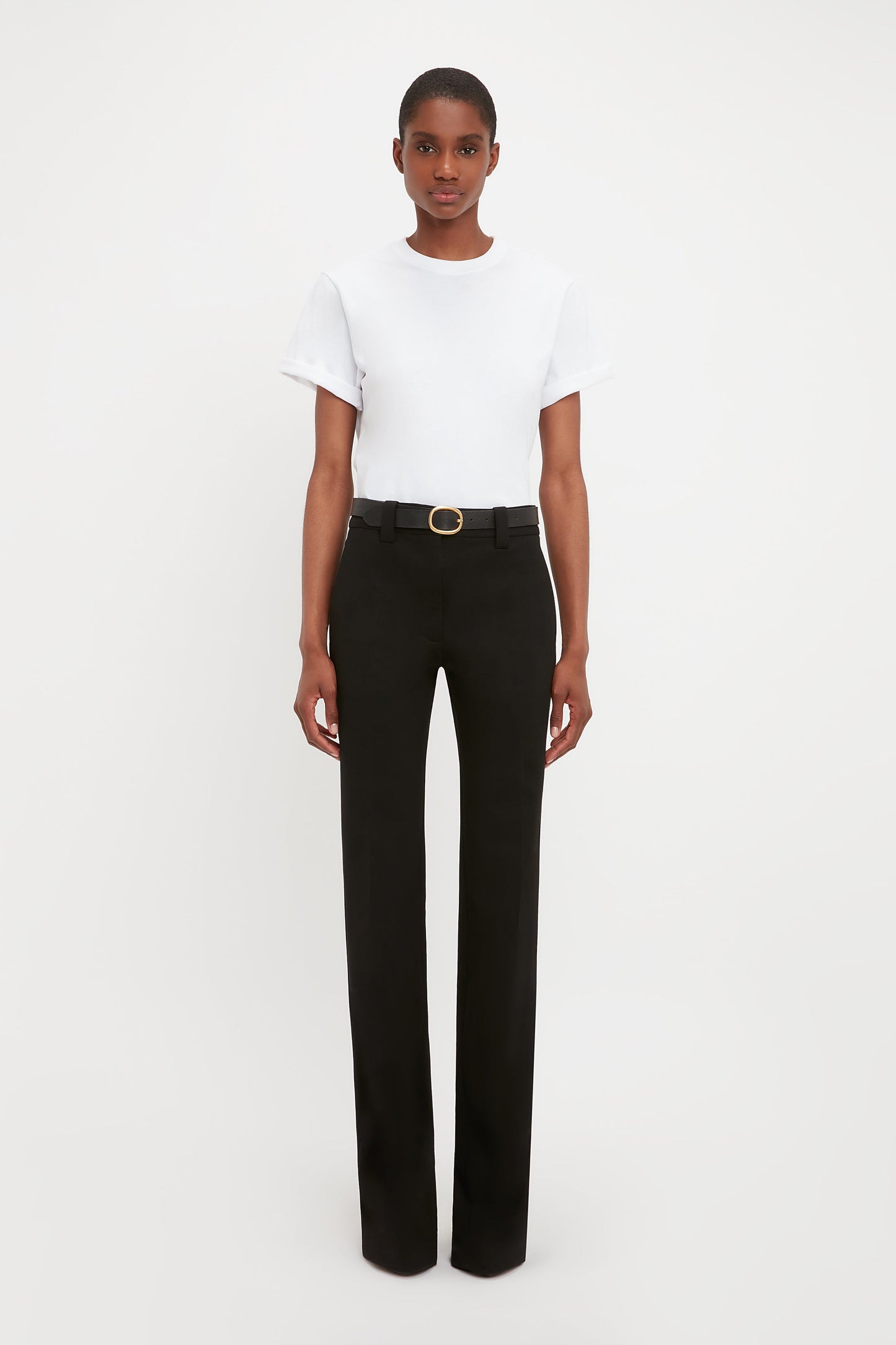 A person stands against a plain white background wearing a white short-sleeve t-shirt, Victoria Beckham Tailored Straight Leg Trouser in Black with a belt, and black shoes.