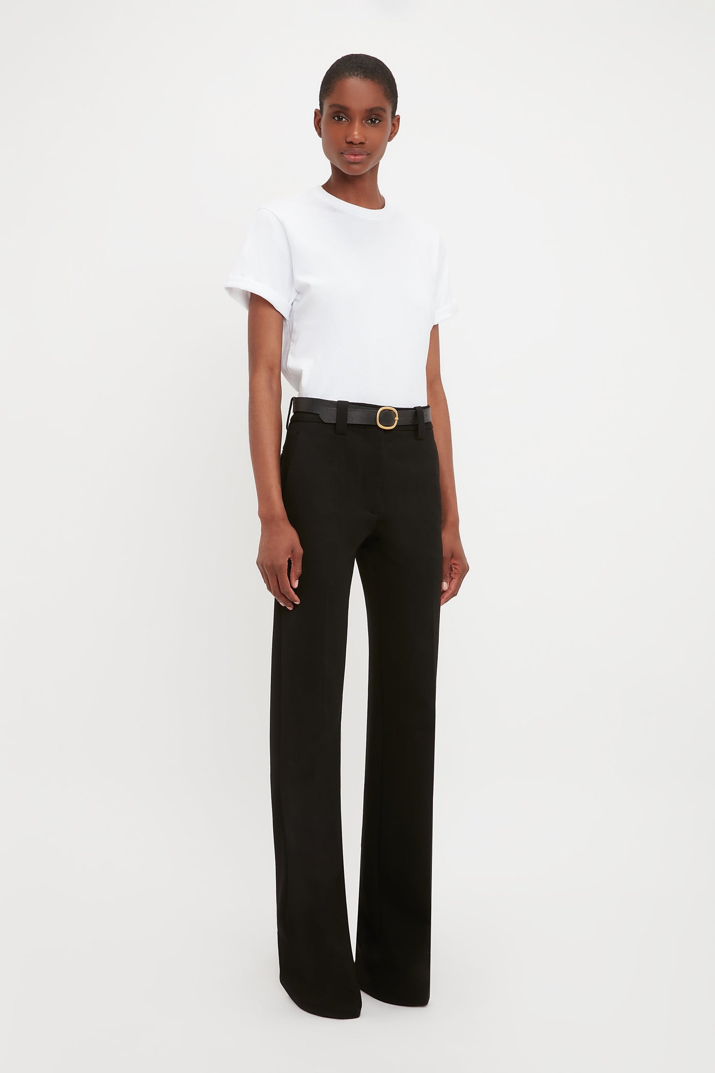 A person stands against a plain white background, wearing Victoria Beckham's Tailored Straight Leg Trouser in Black with a belt and a tucked-in white short-sleeve t-shirt.