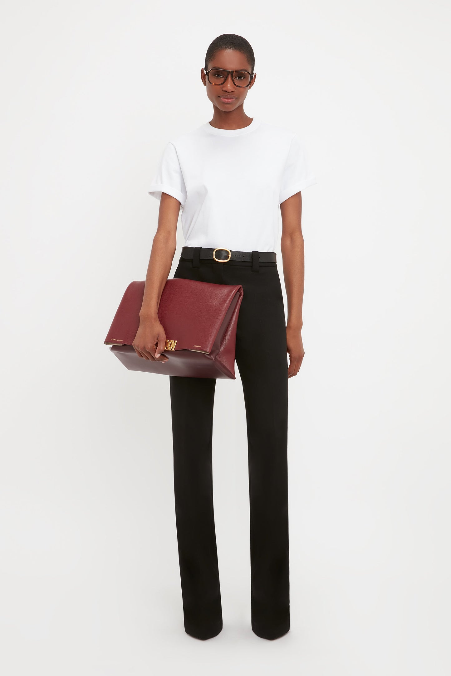 A person wearing a white t-shirt, Victoria Beckham Tailored Straight Leg Trouser In Black with a black belt and gold buckle, glasses, and holding a large dark red portfolio bag against a plain white background.