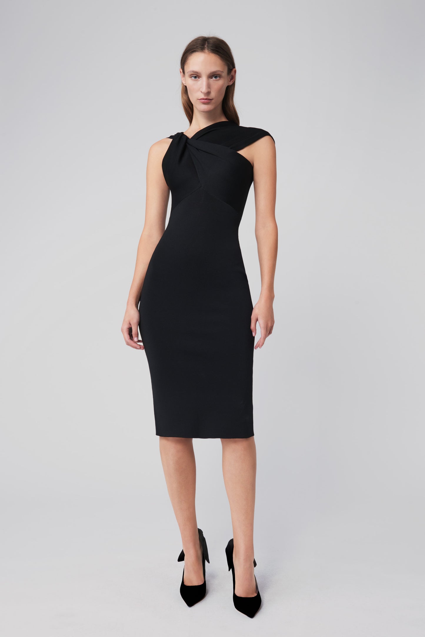 A woman in a black Twist Drape Knit Dress by Victoria Beckham, featuring a form-fitting design and an asymmetric neckline, stands against a light gray background.