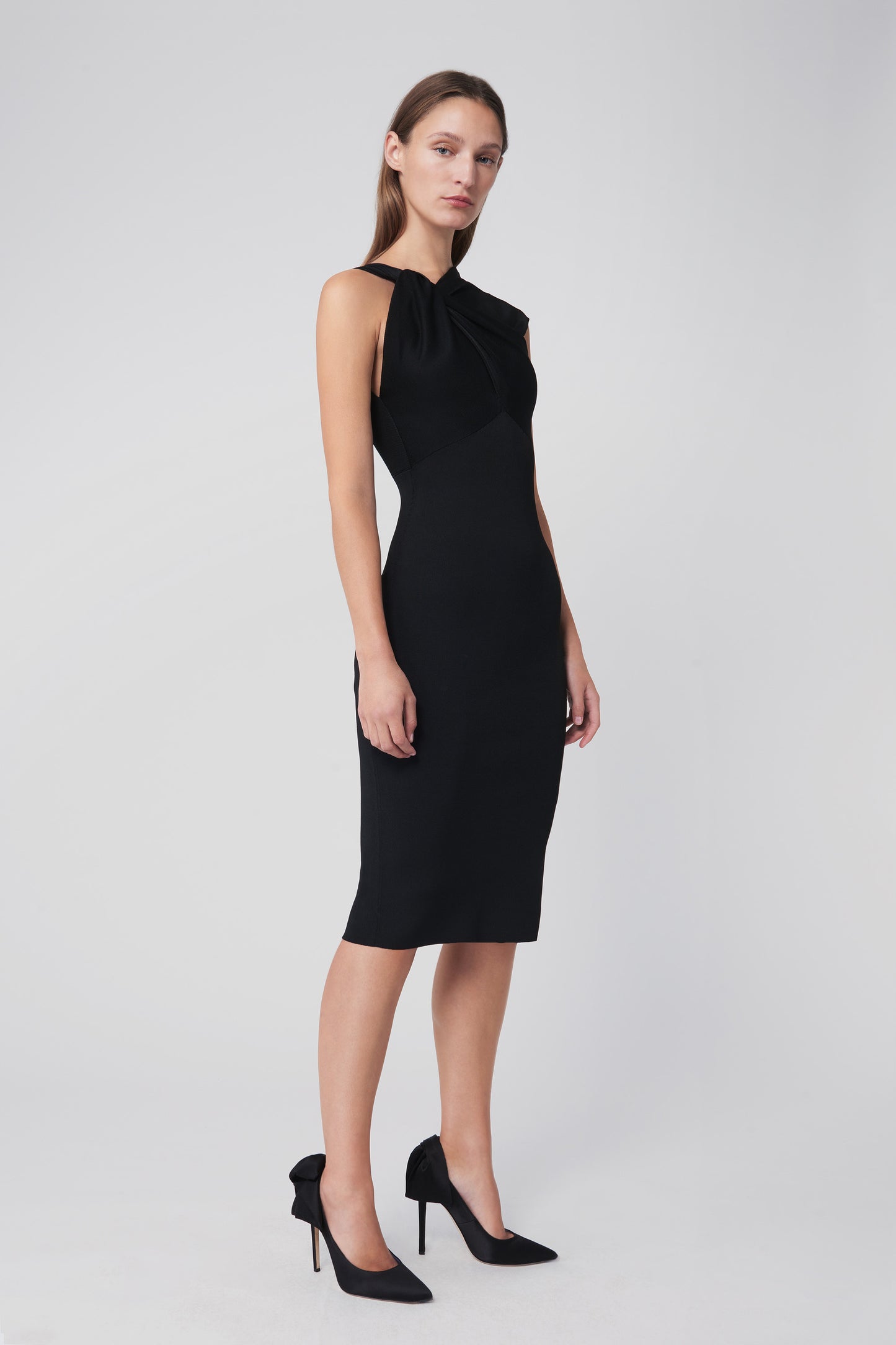 A woman wearing the Victoria Beckham Twist Drape Knit Dress in Black, featuring a sleeveless design and an asymmetric neckline with twist draping, stands against a neutral background, her high heels completing the chic ensemble.